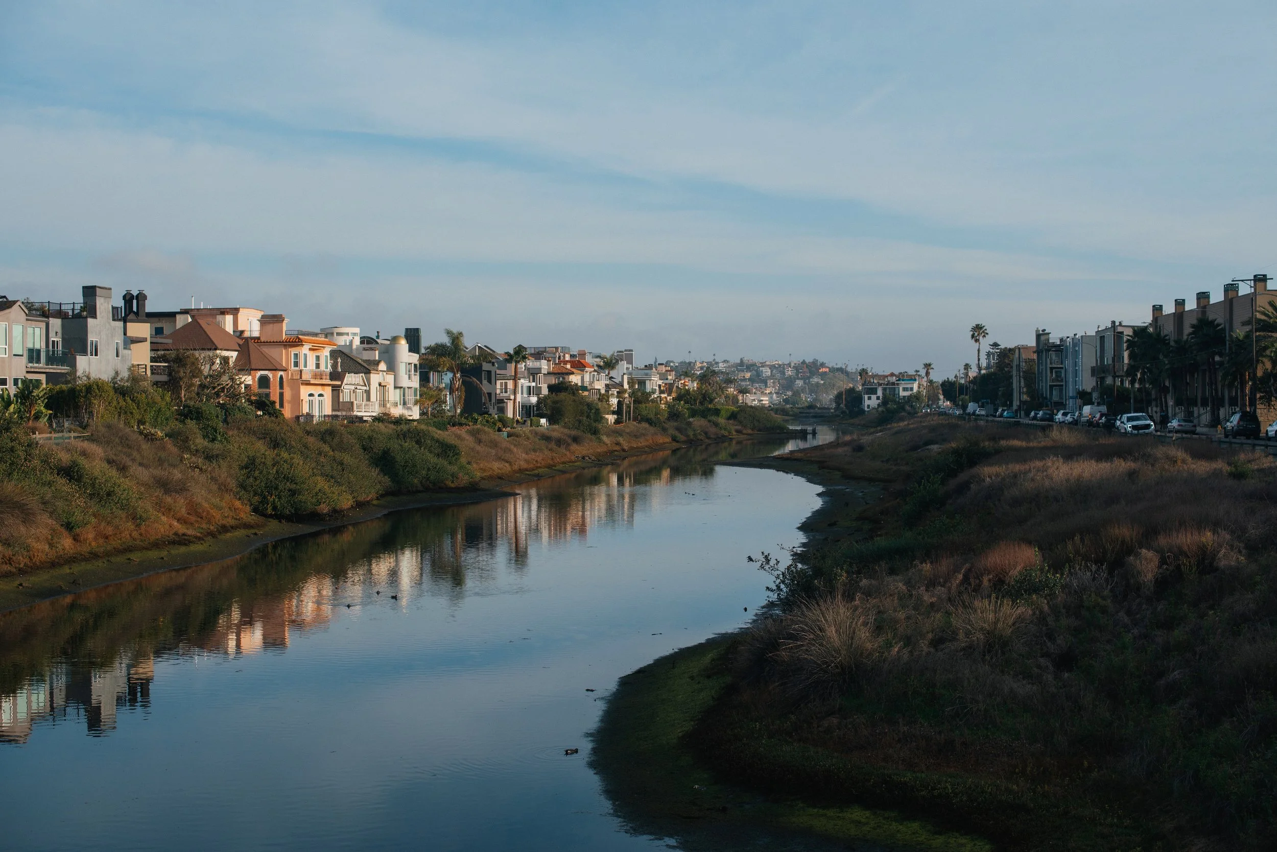 A cityscape showing a creek running through a residential area with houses lined along both sides, with a clear sky overhead and distant hills in the background.