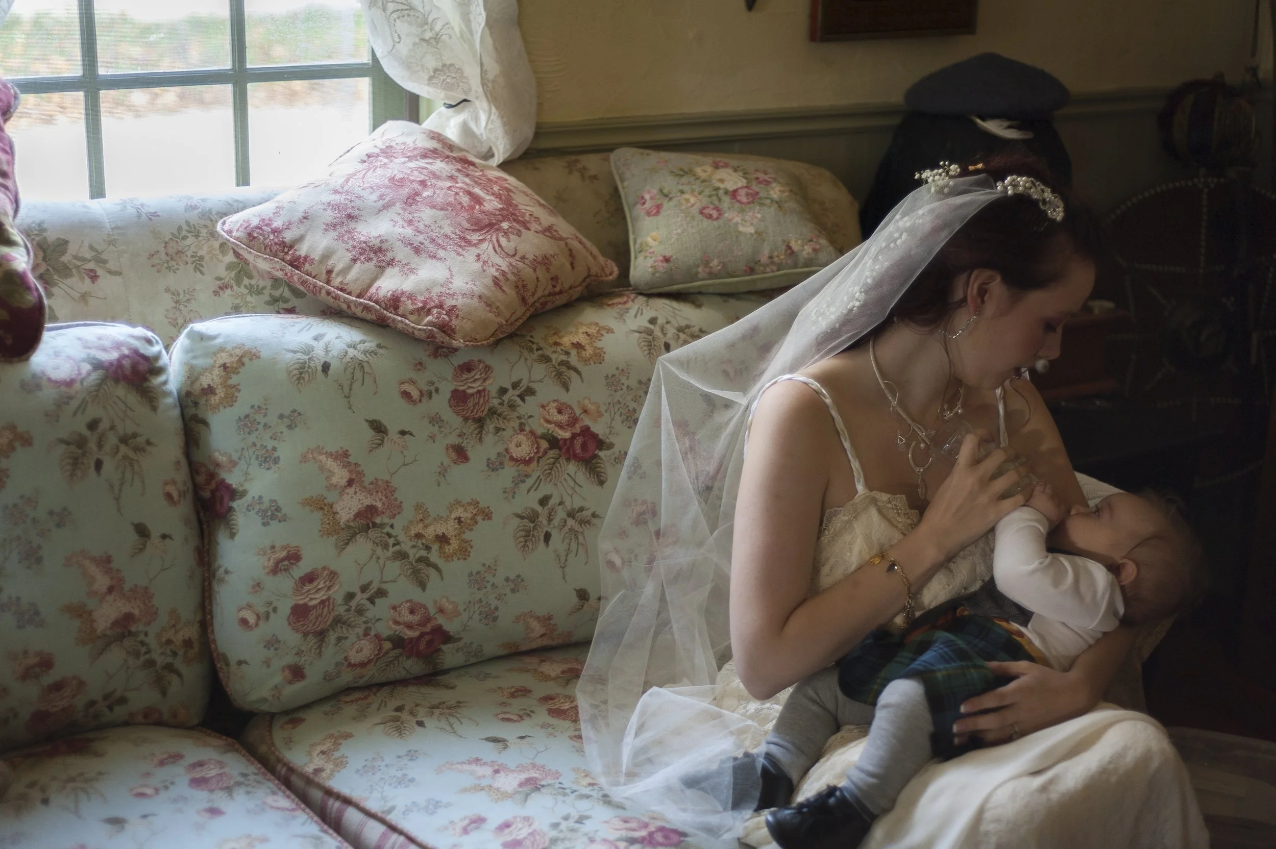 A woman dressed as a bride, wearing a wedding veil and jewelry, sitting on a floral-patterned couch and holding a young boy who is drinking from her.