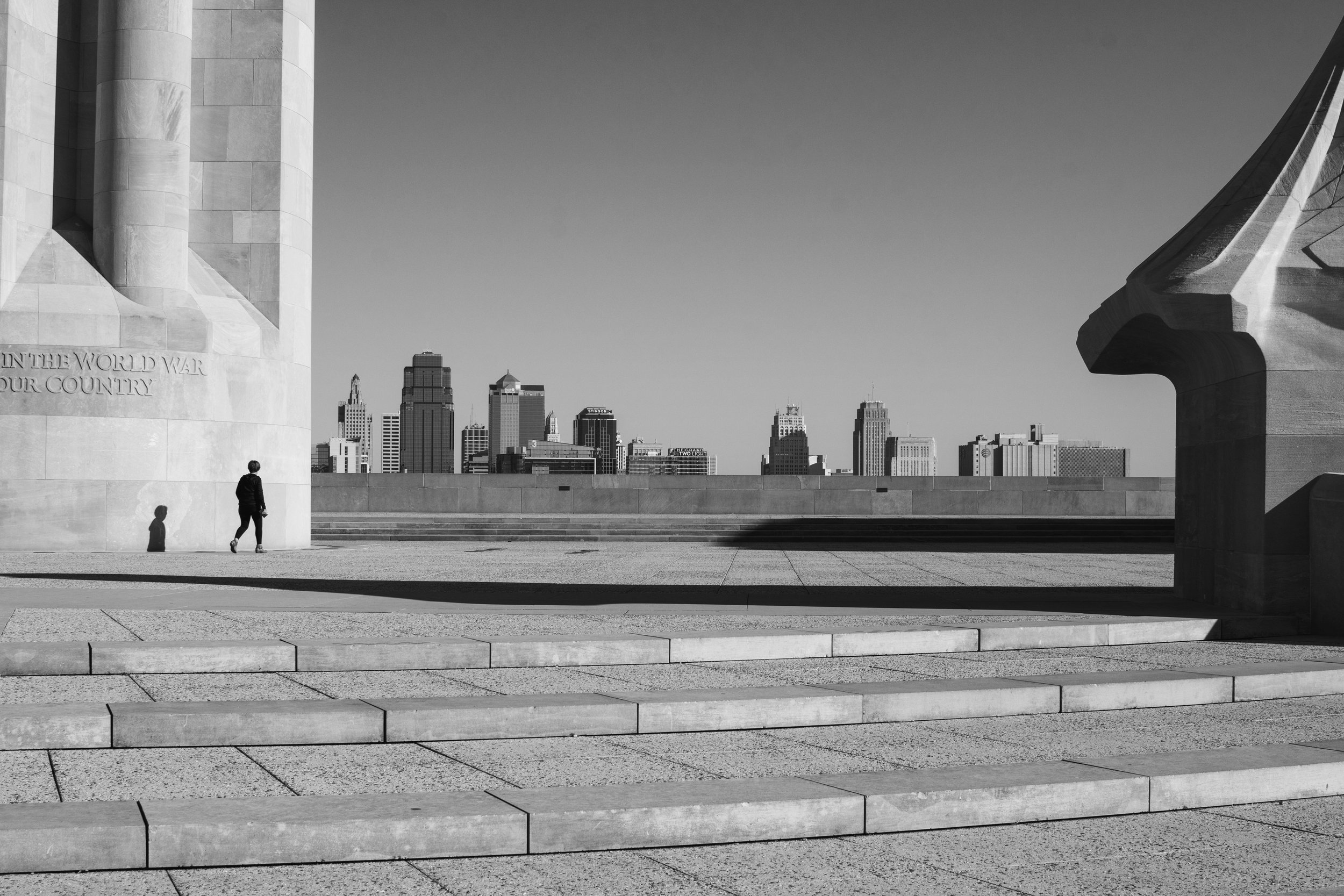 A person walking on an open paved area in front of a large monument with a city skyline in the background, in black and white.