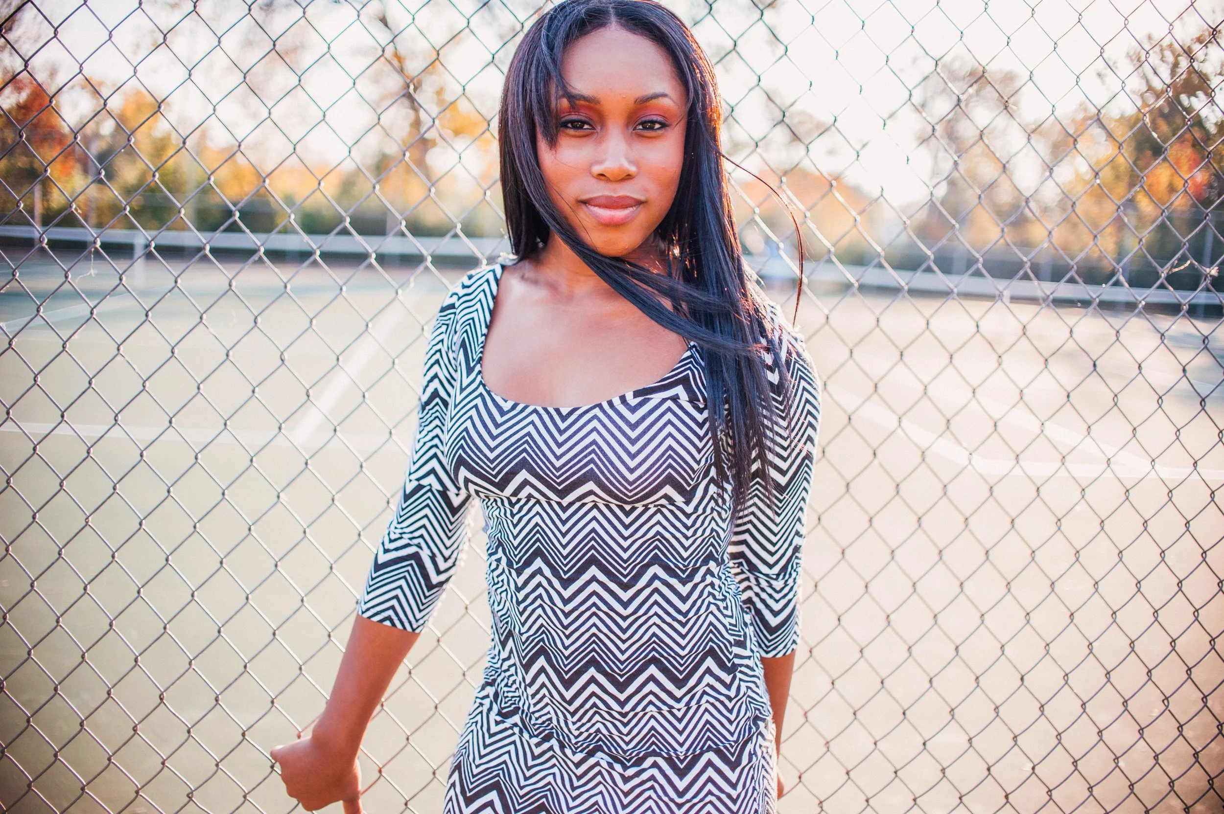 A woman with long black hair wearing a black and white zigzag patterned dress standing in front of a metal chain-link fence at an outdoor tennis or sports court during sunset.