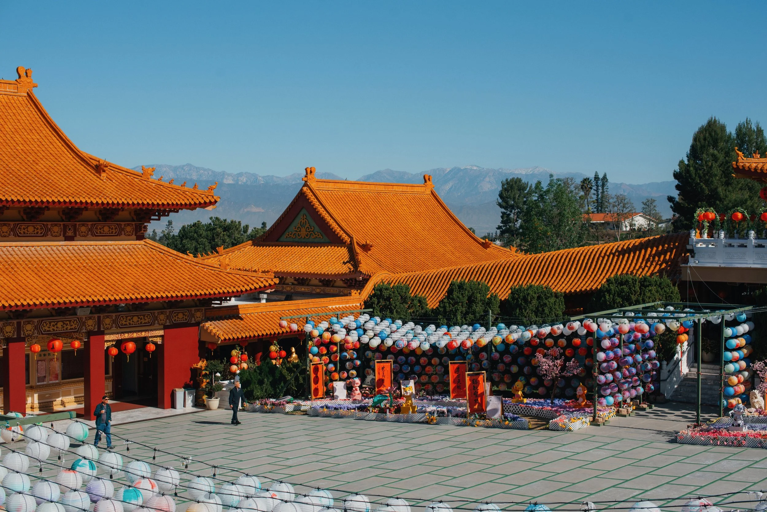 A traditional Chinese temple with orange roofs decorated with lanterns and banners, set against a backdrop of mountains and trees.
