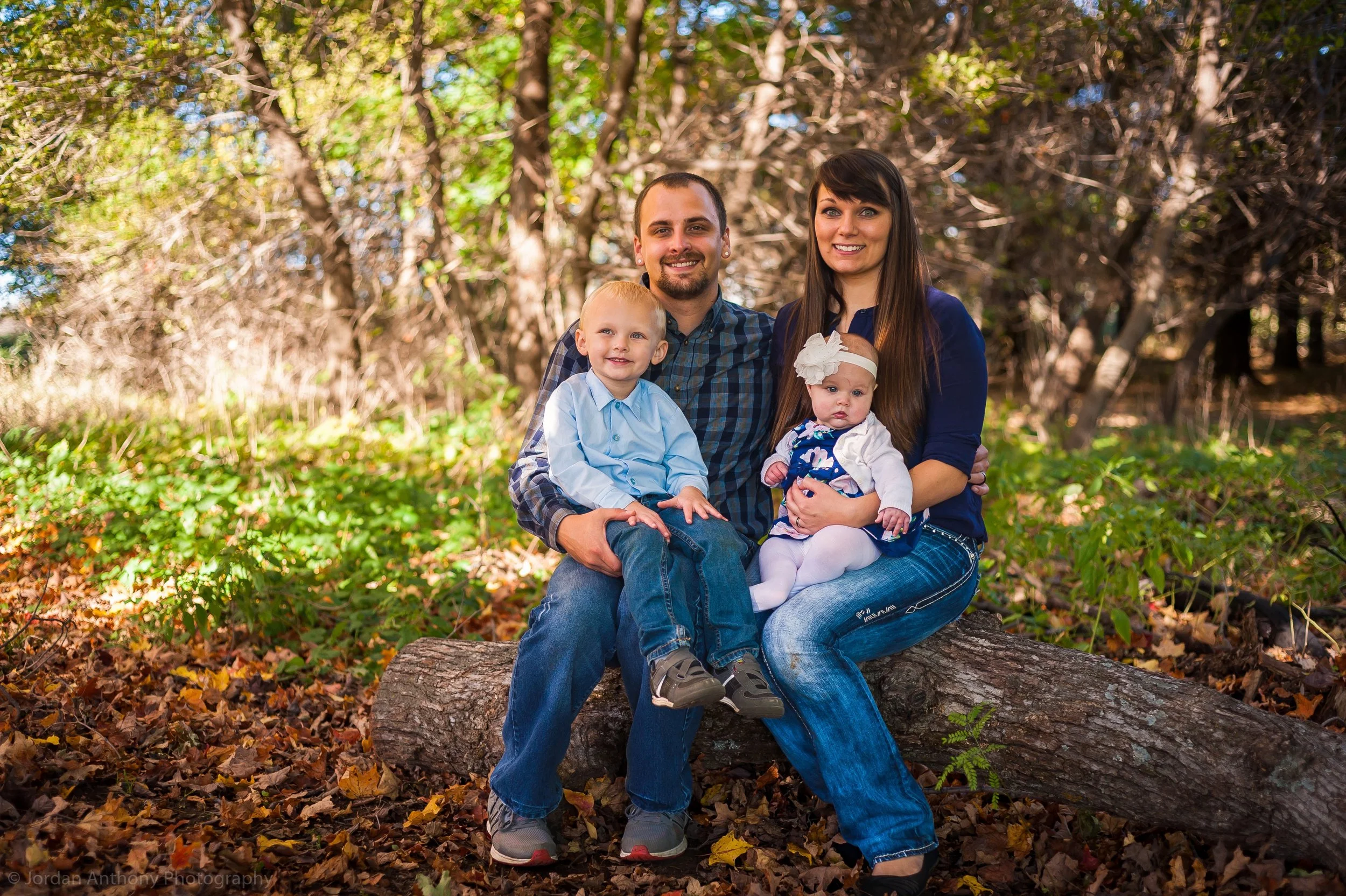 A family of four sitting on a fallen log in a wooded area during fall, smiling at the camera. The father and older son are on the left, the mother with the baby girl on her lap are on the right.