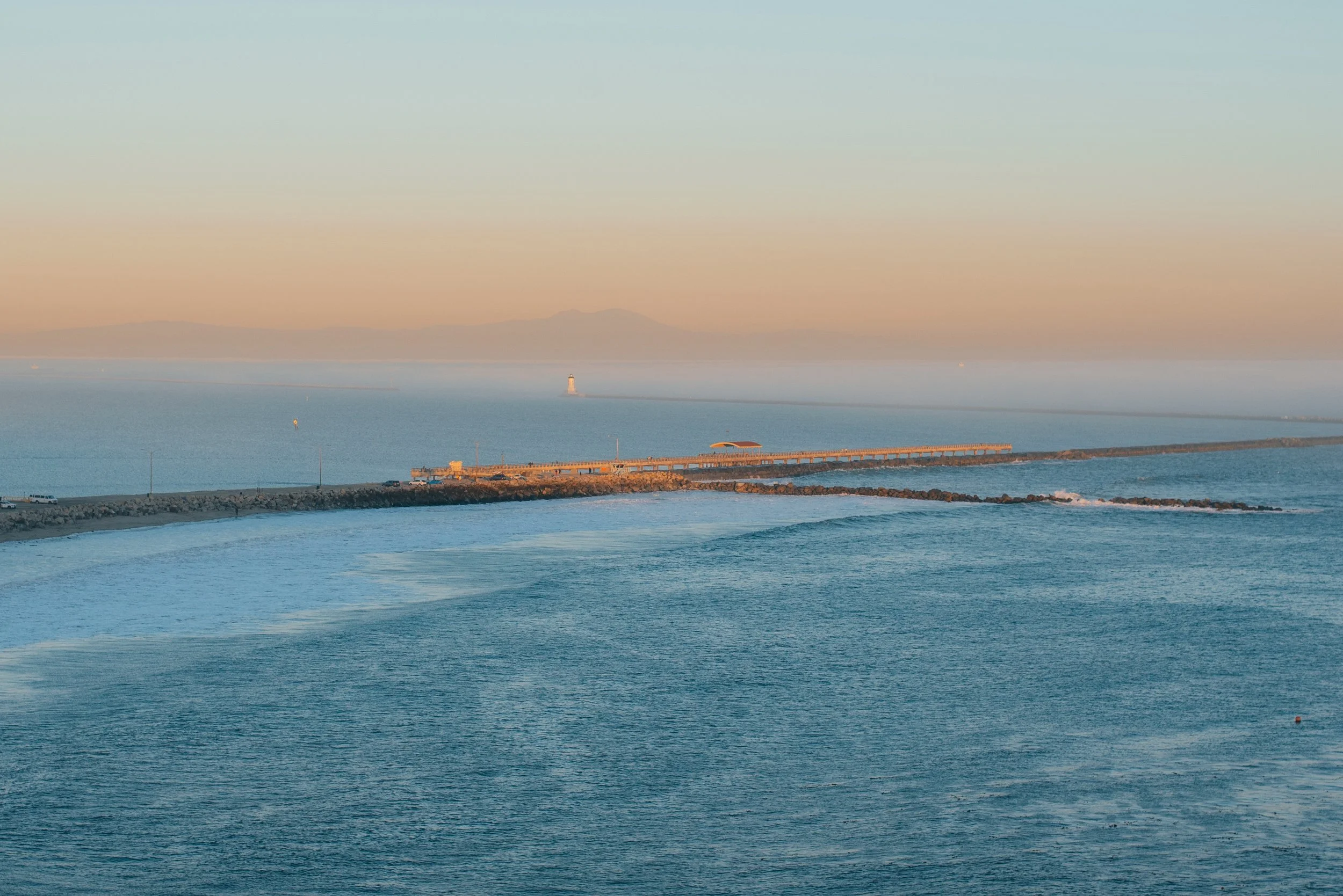 View of the ocean with a breakwater extending into the water and a lighthouse in the distance during sunset.
