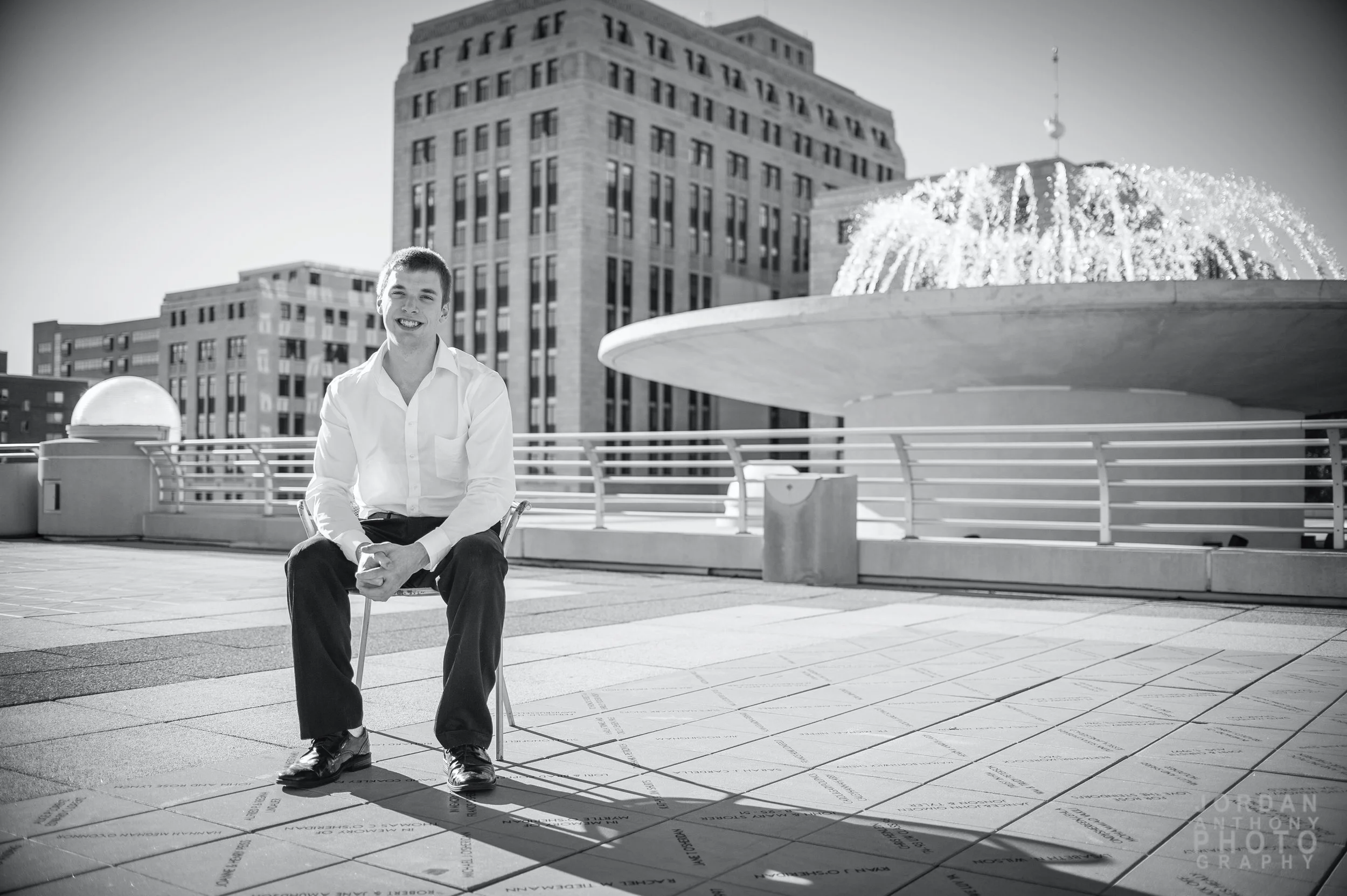 A young man in a white dress shirt and black pants sitting on a chair on an urban rooftop with skyscrapers and a fountain in the background.