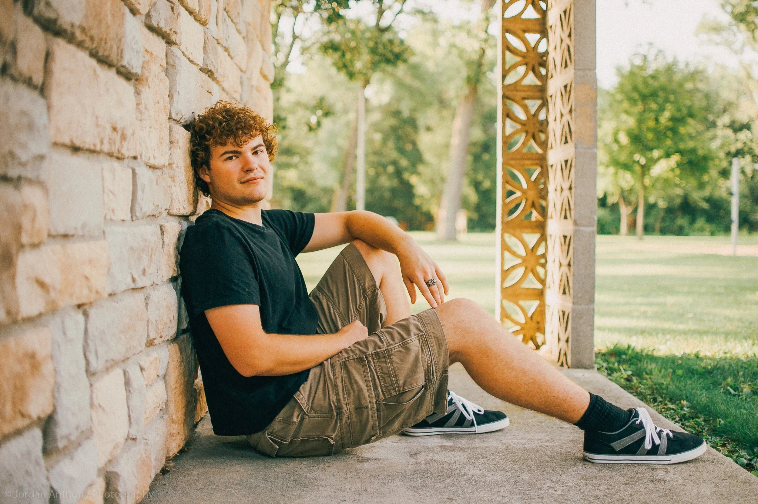 A young man with curly hair sitting on the ground, leaning against a brick wall on the left. He is wearing a black t-shirt, khaki shorts, and black sneakers with white laces. The background features a park with trees and a patterned lattice structure