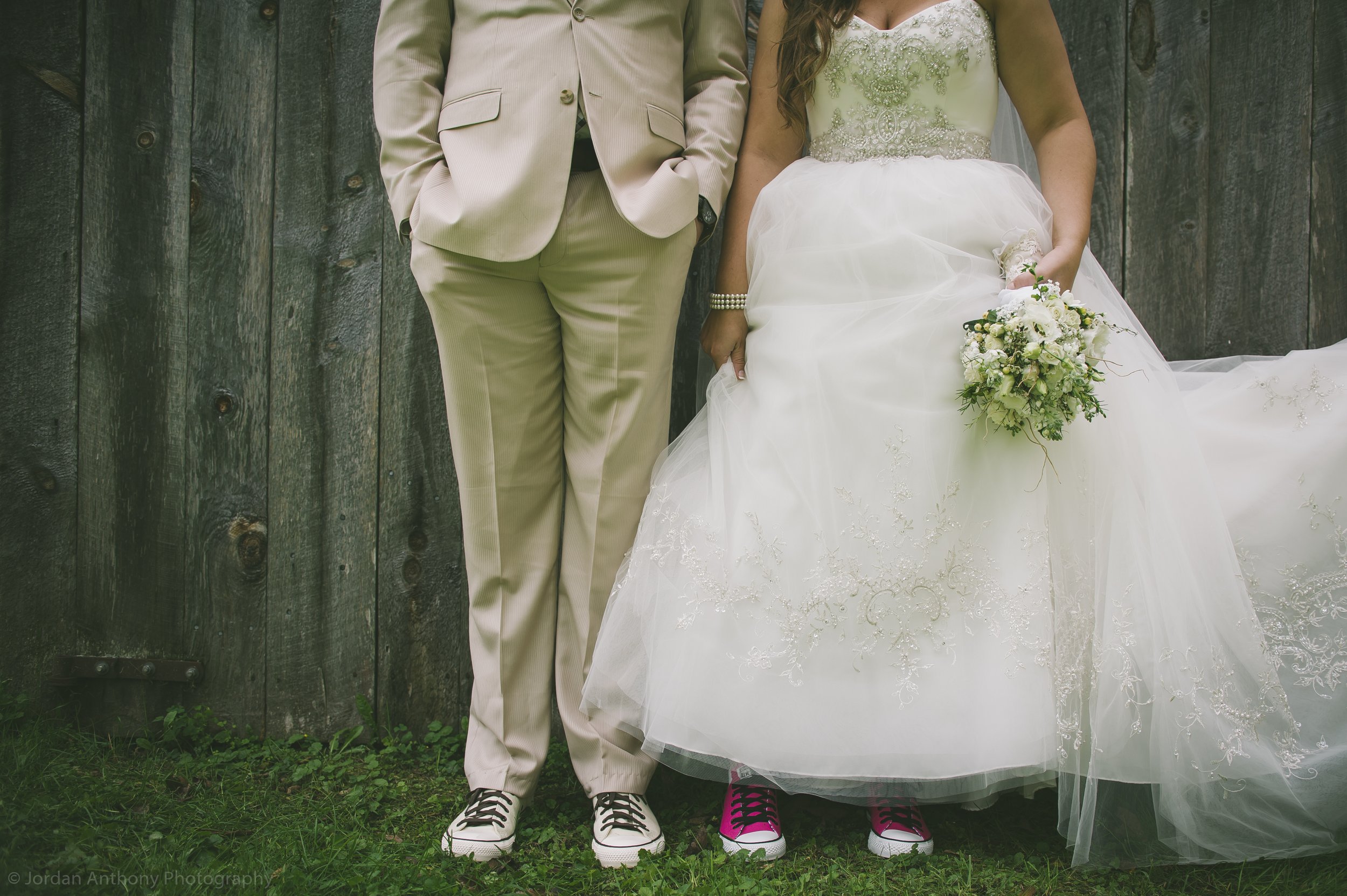 A couple in wedding attire, with the groom in a beige suit and the bride in a white wedding dress, standing against a wooden fence. Both are wearing colorful sneakers and holding a bouquet.