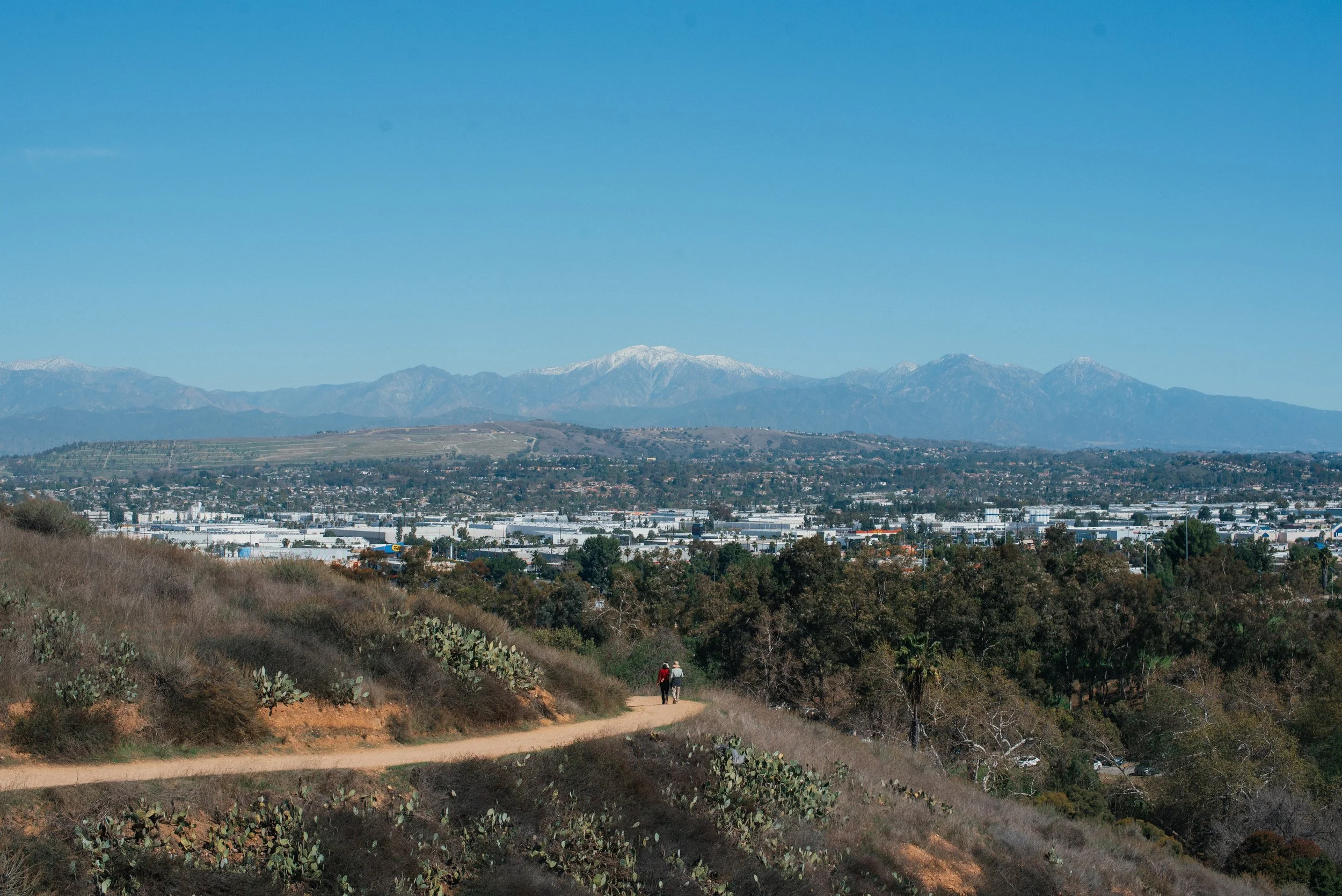 Hiking trail on a hillside with two people walking, cityscape with industrial buildings and trees in the middle ground, or mountains with snow in the background under a clear blue sky.