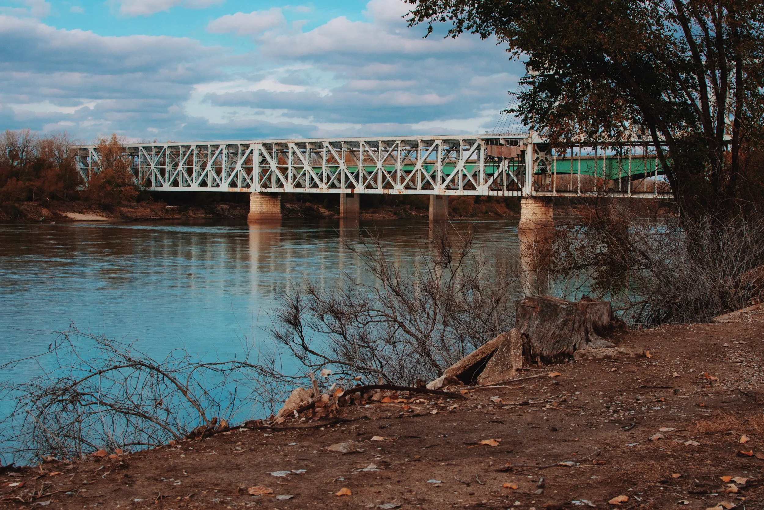 View of a metal bridge over a river with trees and cloudy sky in the background.