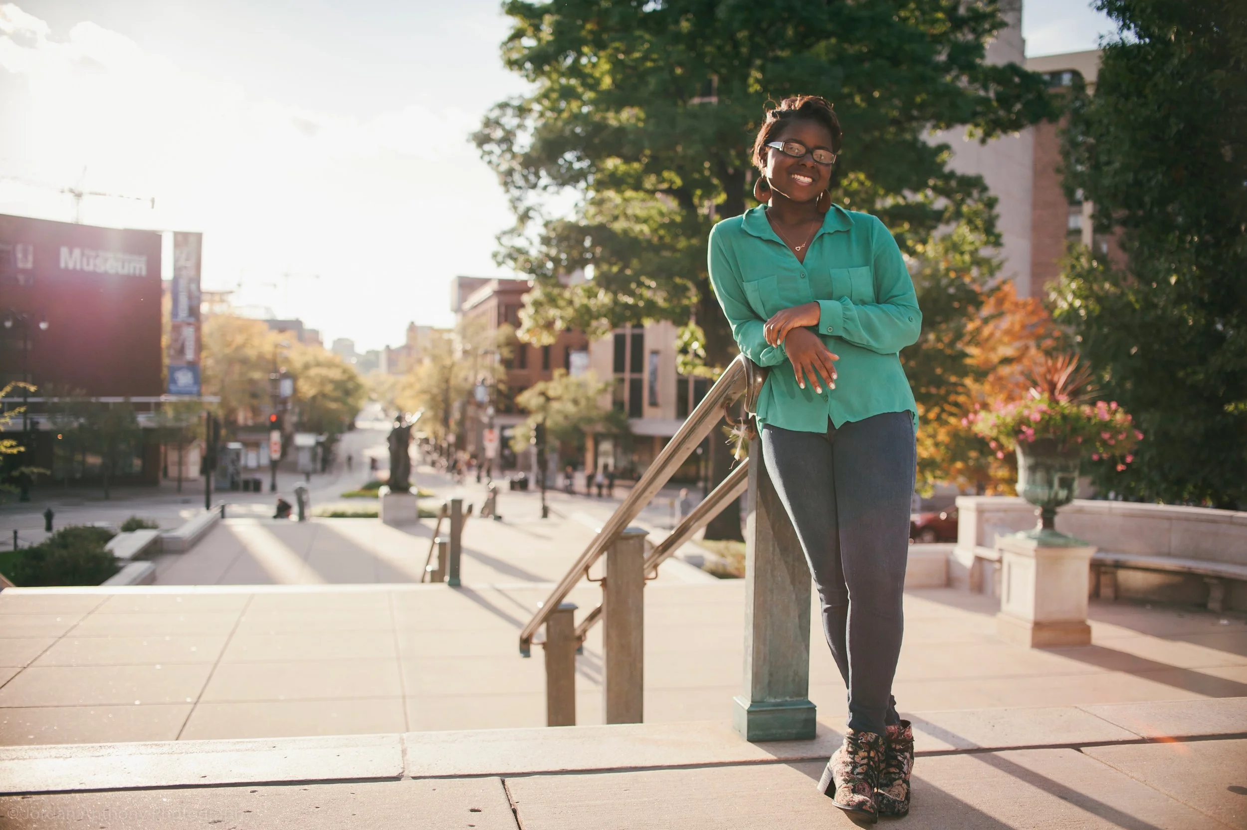 A woman with glasses wearing a turquoise shirt and dark jeans standing on a city street steps, smiling with arms crossed, with trees and buildings in the background during sunset.