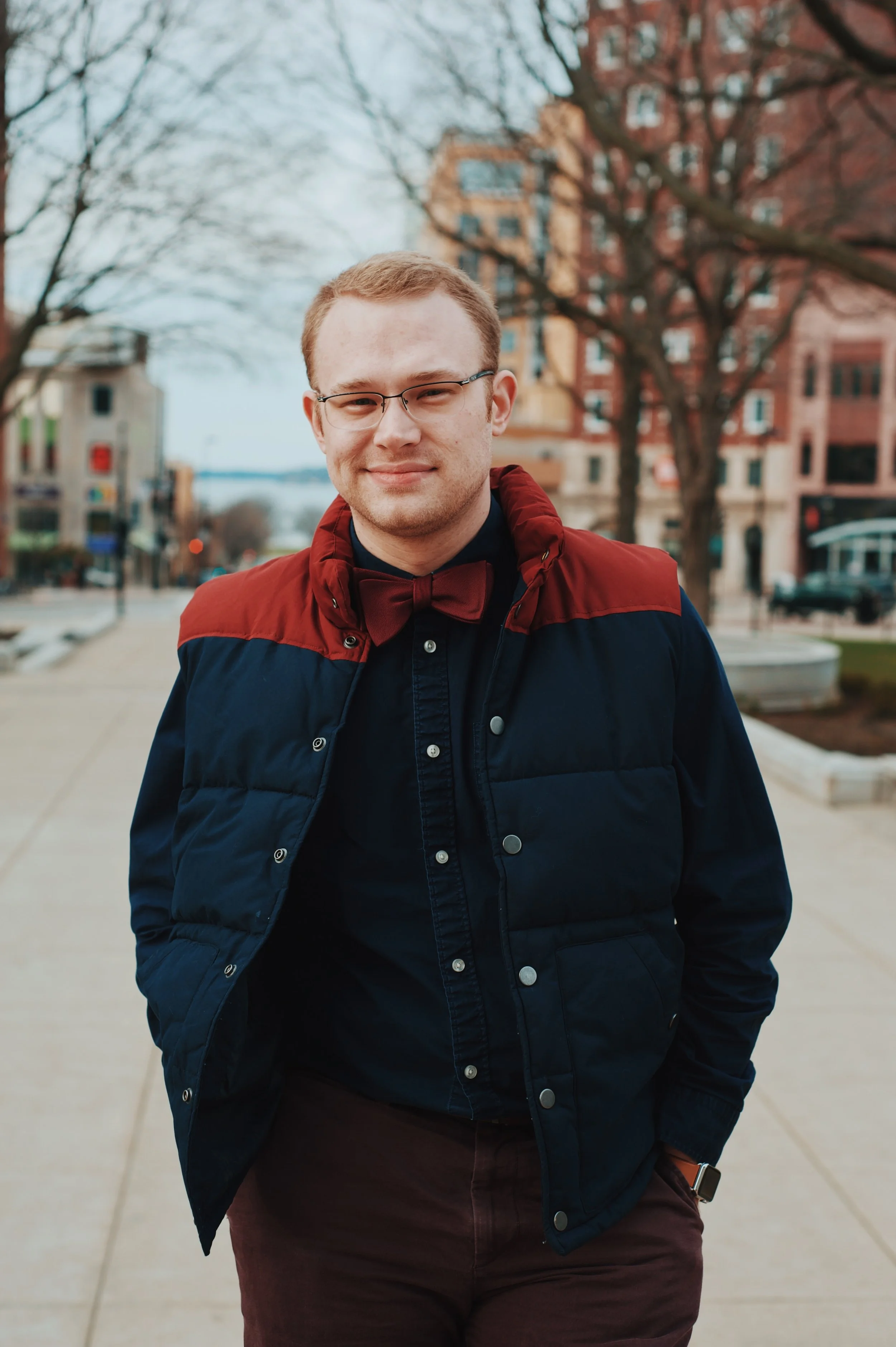A young man with glasses and light brown hair, standing outdoors on a sidewalk in front of trees and buildings during daytime, wearing a navy blue and red jacket, a maroon bow tie, and a smartwatch.