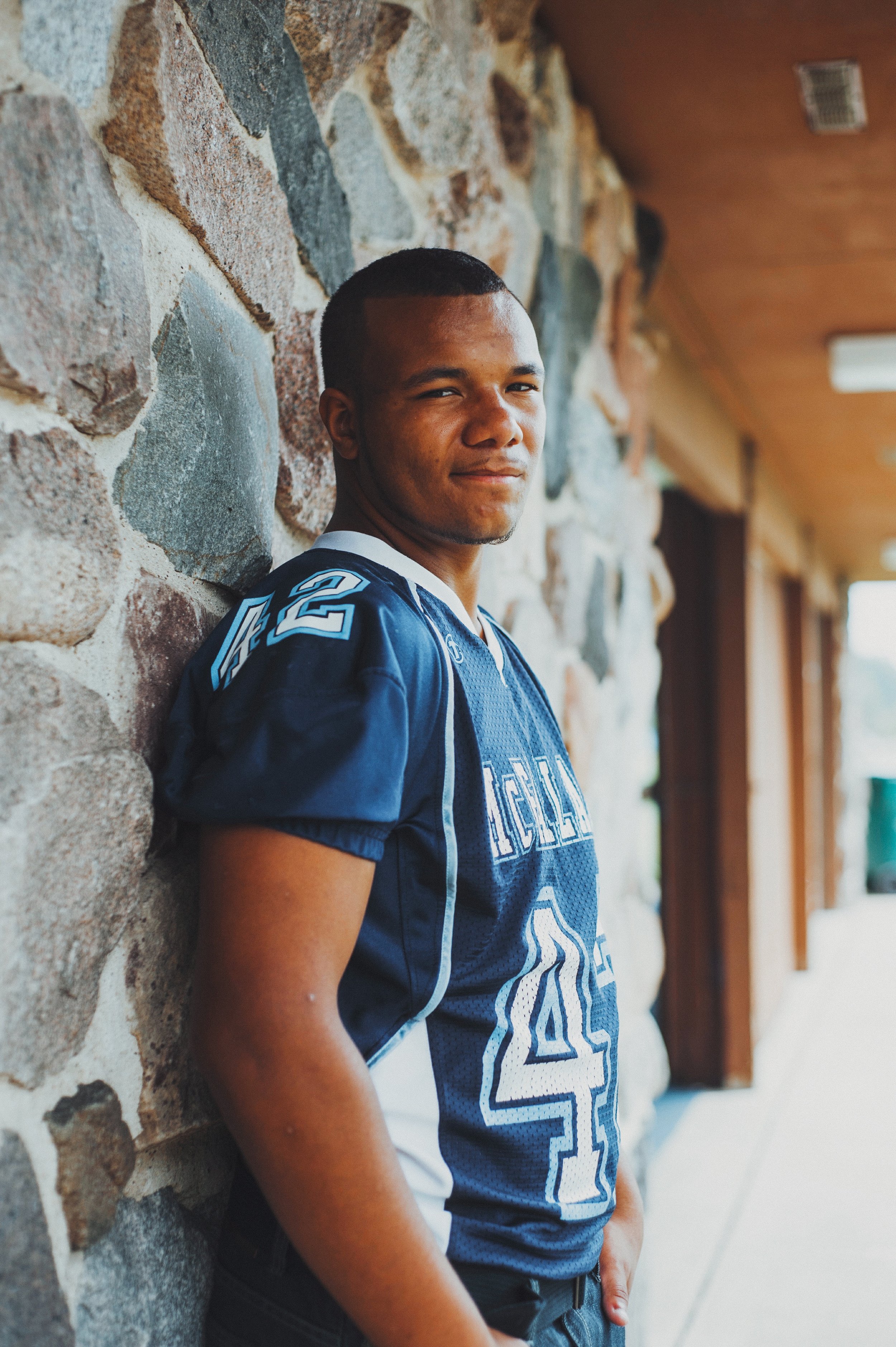 Young man in a blue football jersey leaning against a stone wall, looking at the camera with a slight smile.