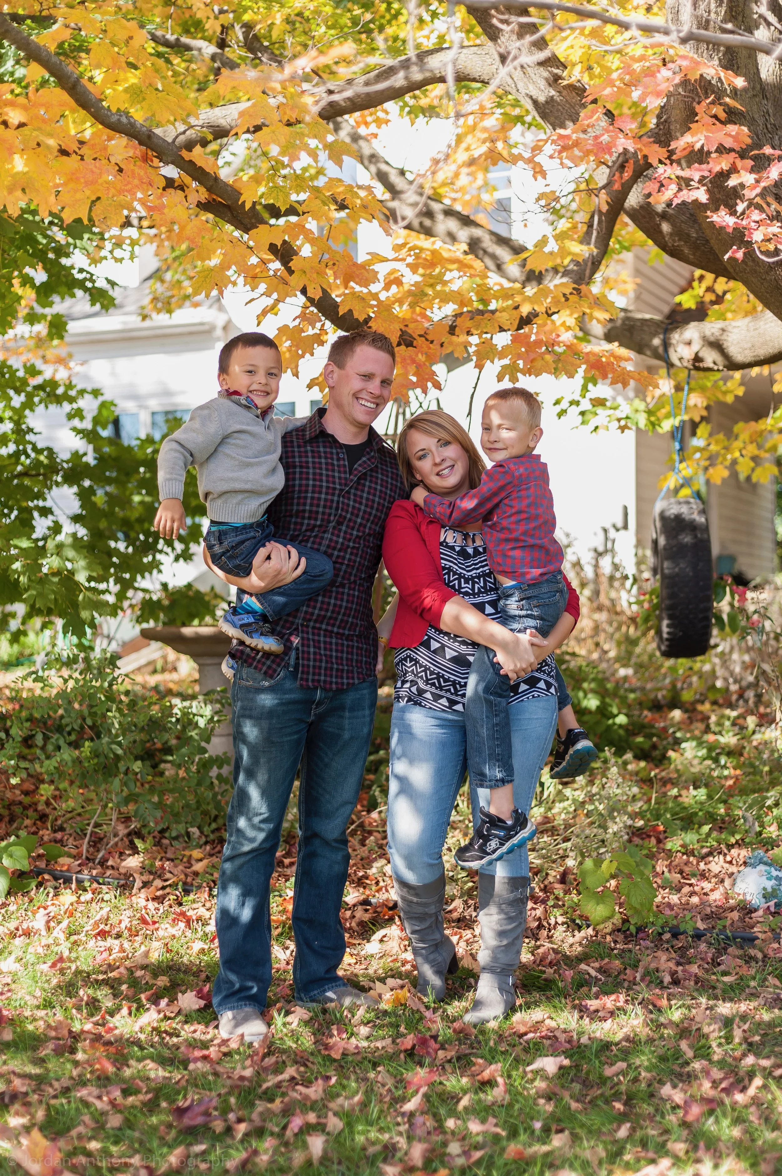 A family of four smiling and standing outdoors on a fall day, with fallen leaves on the ground and orange and yellow leaves on the trees. The father is holding a young boy, and the mother is holding another child, with a tire swing hanging from a tre
