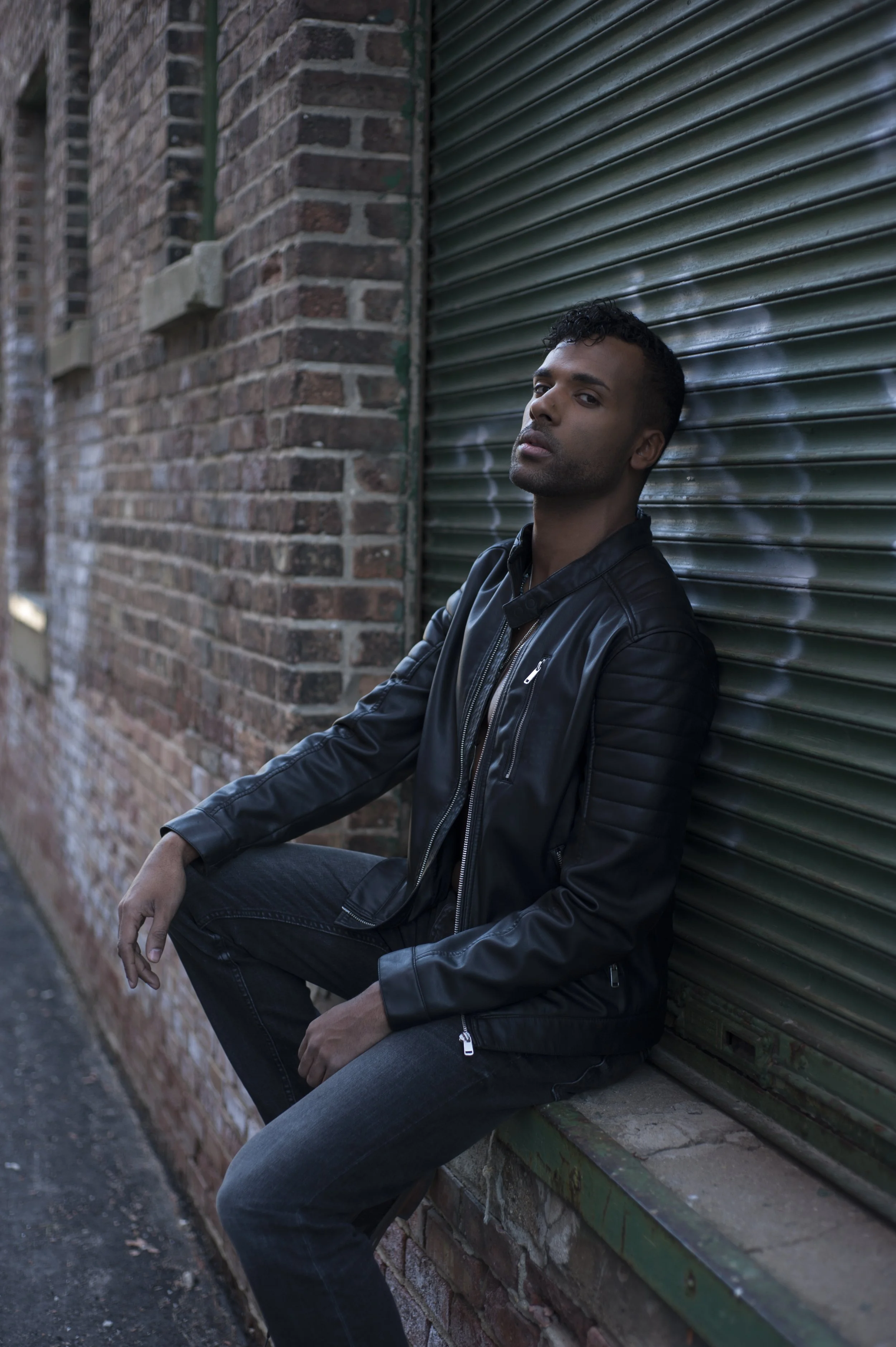 A young man sitting on a brick ledge against a green metal shutter in an urban alleyway, wearing a black leather jacket and jeans.