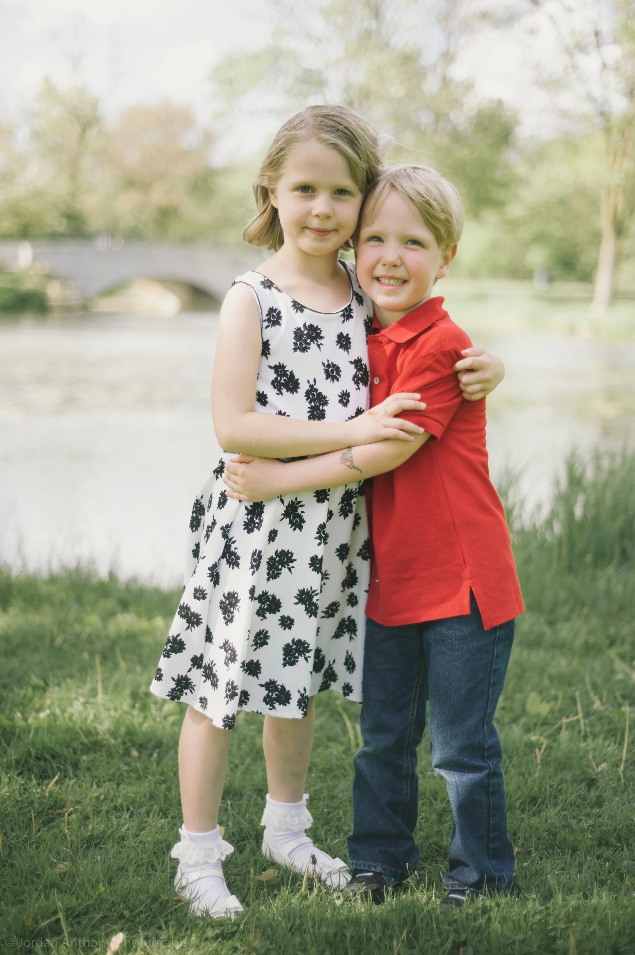 A boy and girl hugging outdoors near a lake, with trees and a bridge in the background.