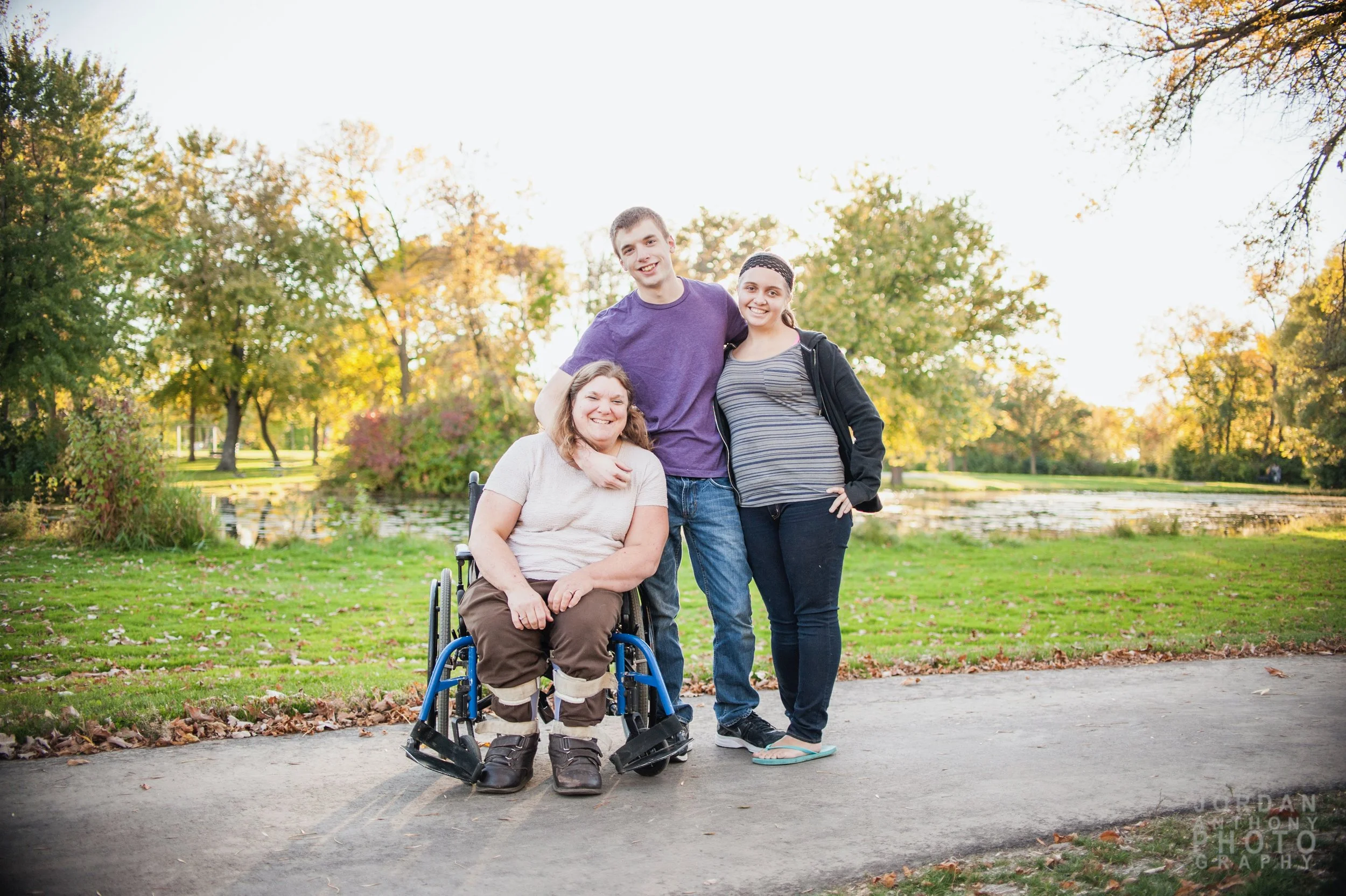 Three people smiling outdoors in a park with trees and a pond, one in a wheelchair, during fall.