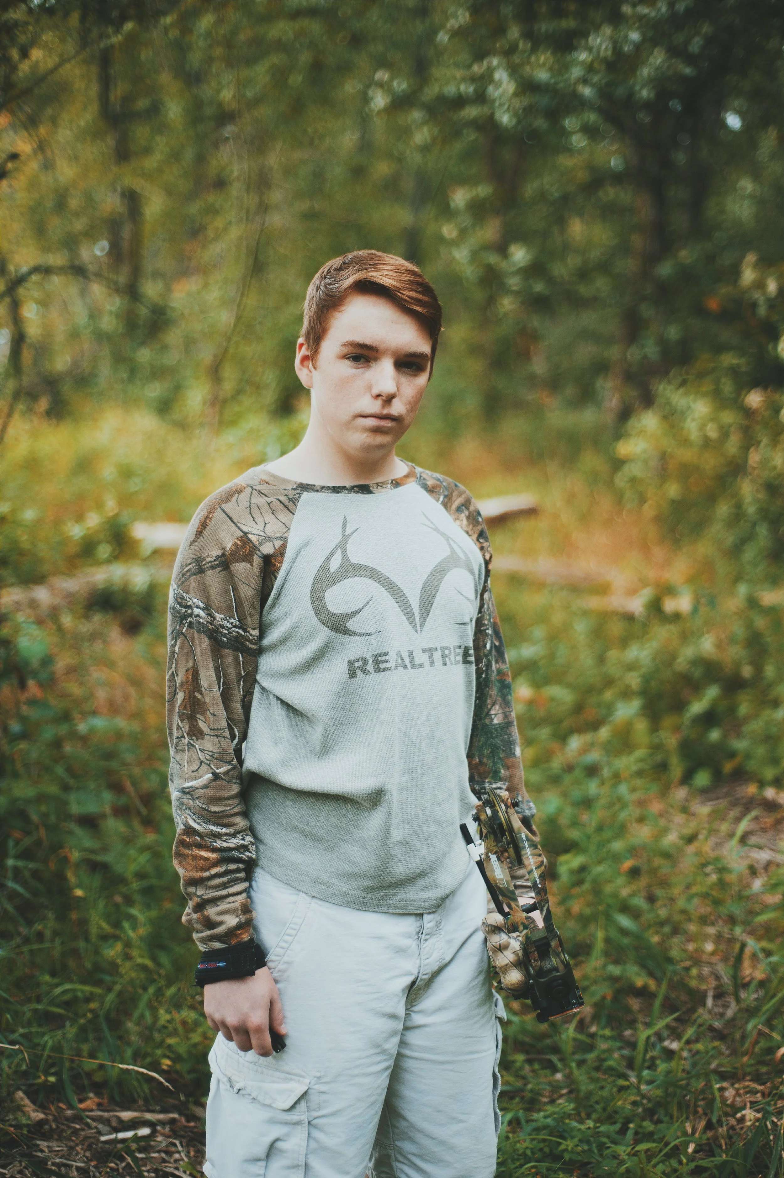 Young man standing outdoors in a forest during fall, wearing a camouflage long sleeve shirt and white cargo shorts, holding a camouflage rifle.