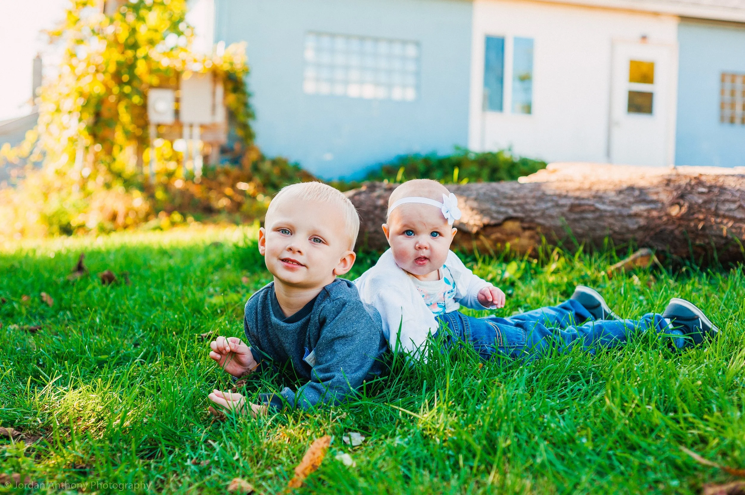 Two young children, a boy and a girl, lying on green grass in a backyard with a fallen log and a white house in the background.