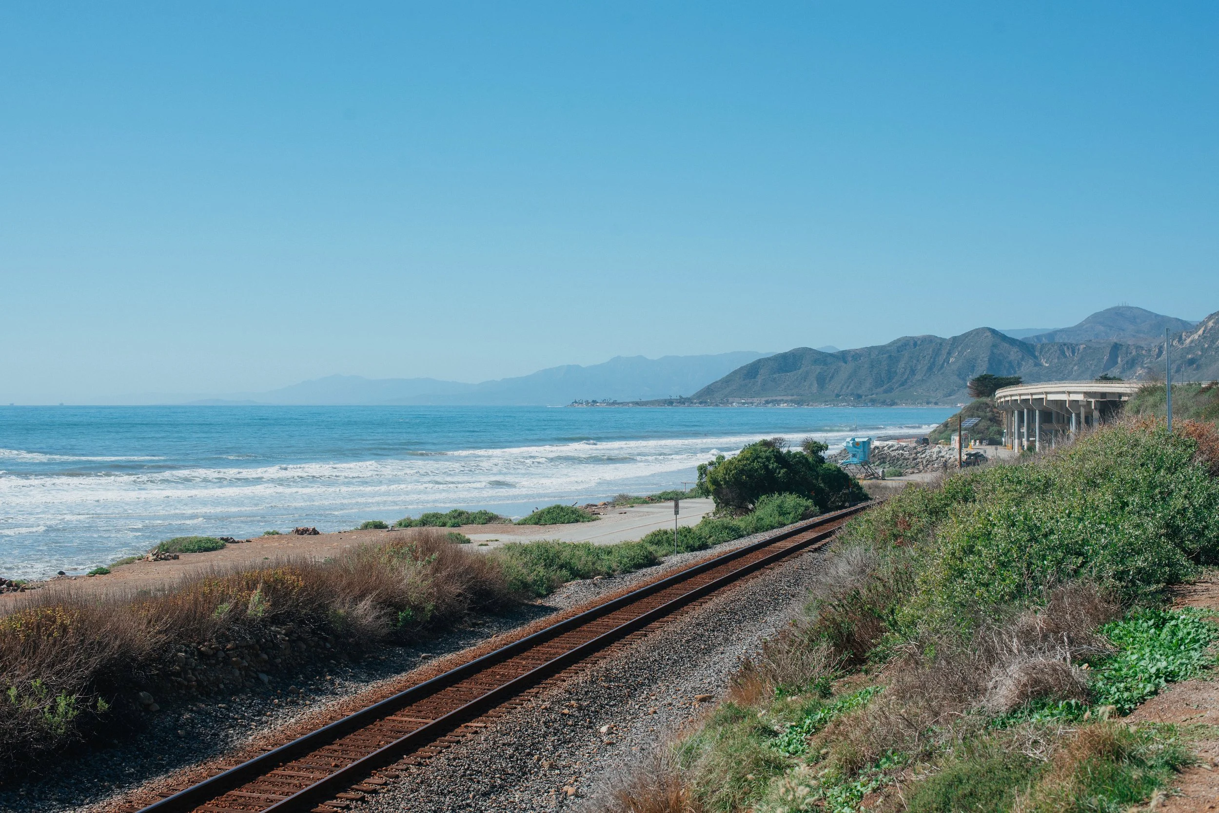 A coastal scene with a train track running parallel to the beach. The ocean waves are crashing on the shore, and there are mountains in the background under a clear blue sky.