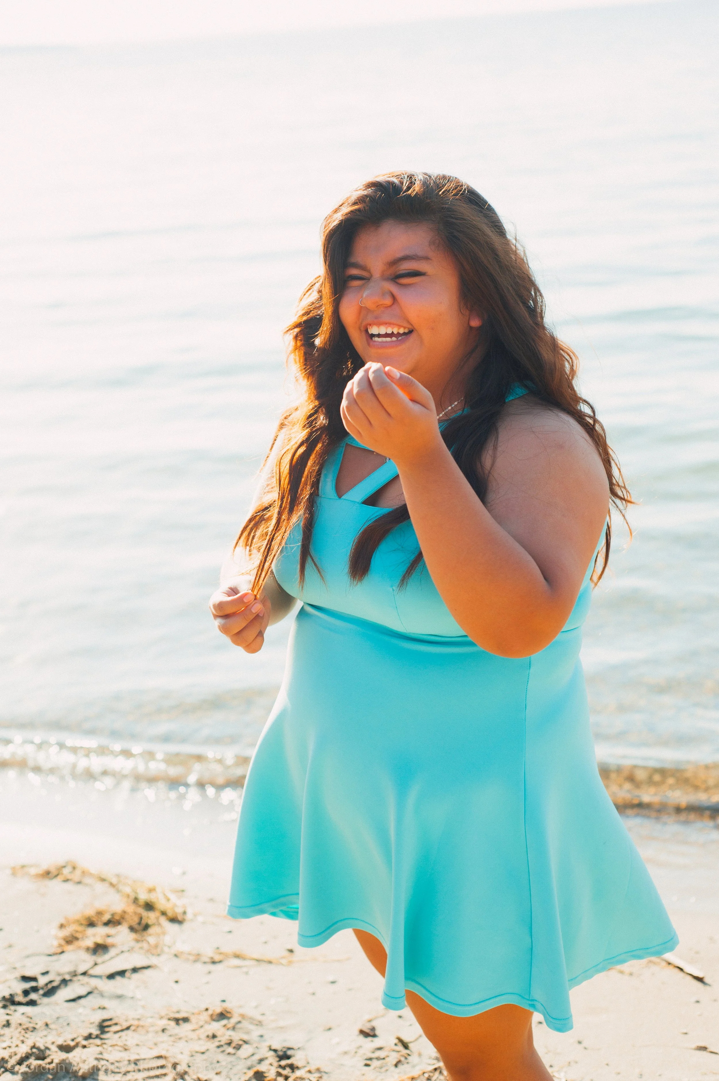 A woman in a light blue dress standing on a sandy beach, smiling and holding a strand of her hair, with the ocean in the background.
