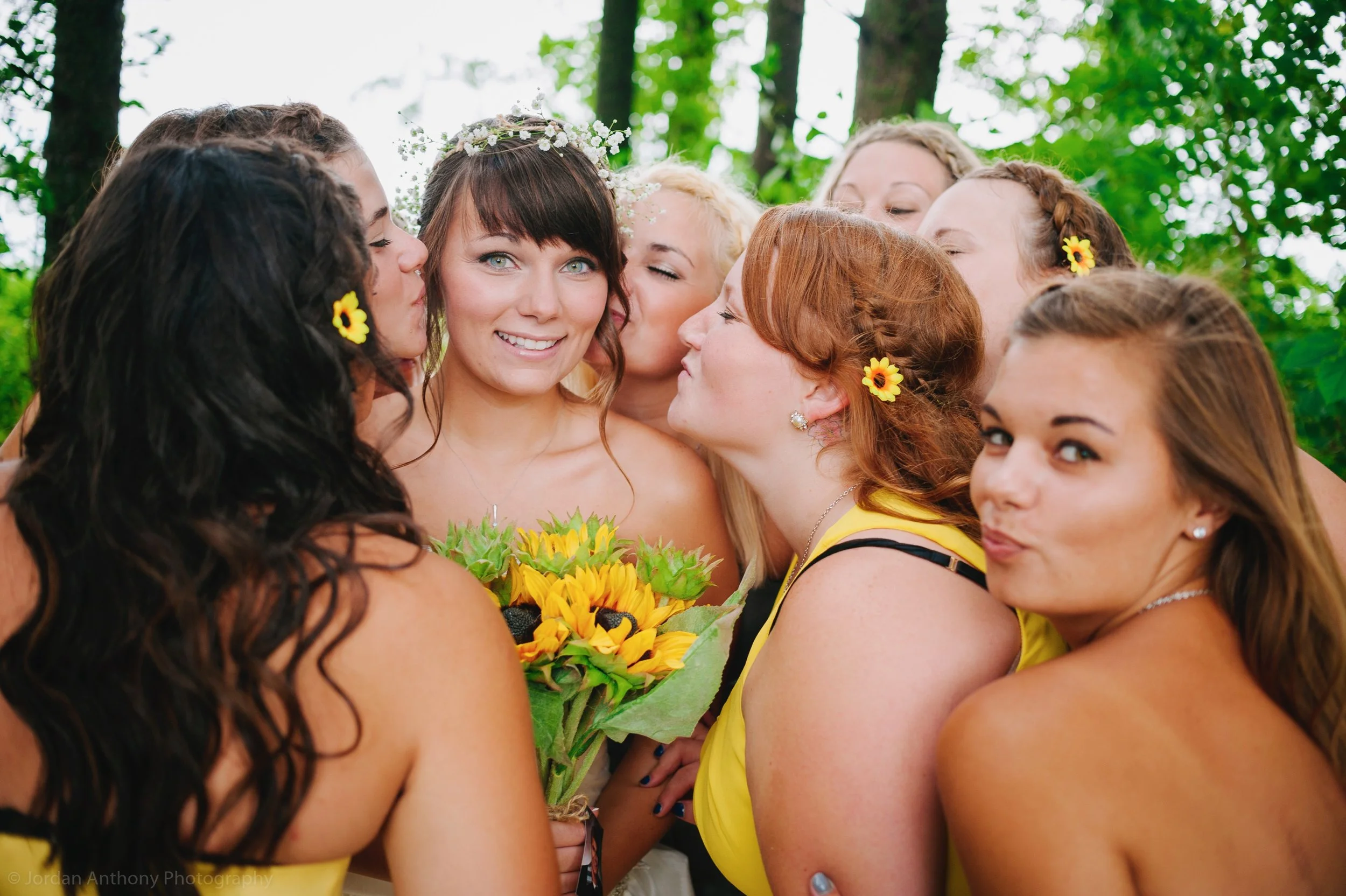 Group of women, some with flowers in their hair, gathered outdoors in a forested area for a celebration, with one holding a sunflower bouquet.