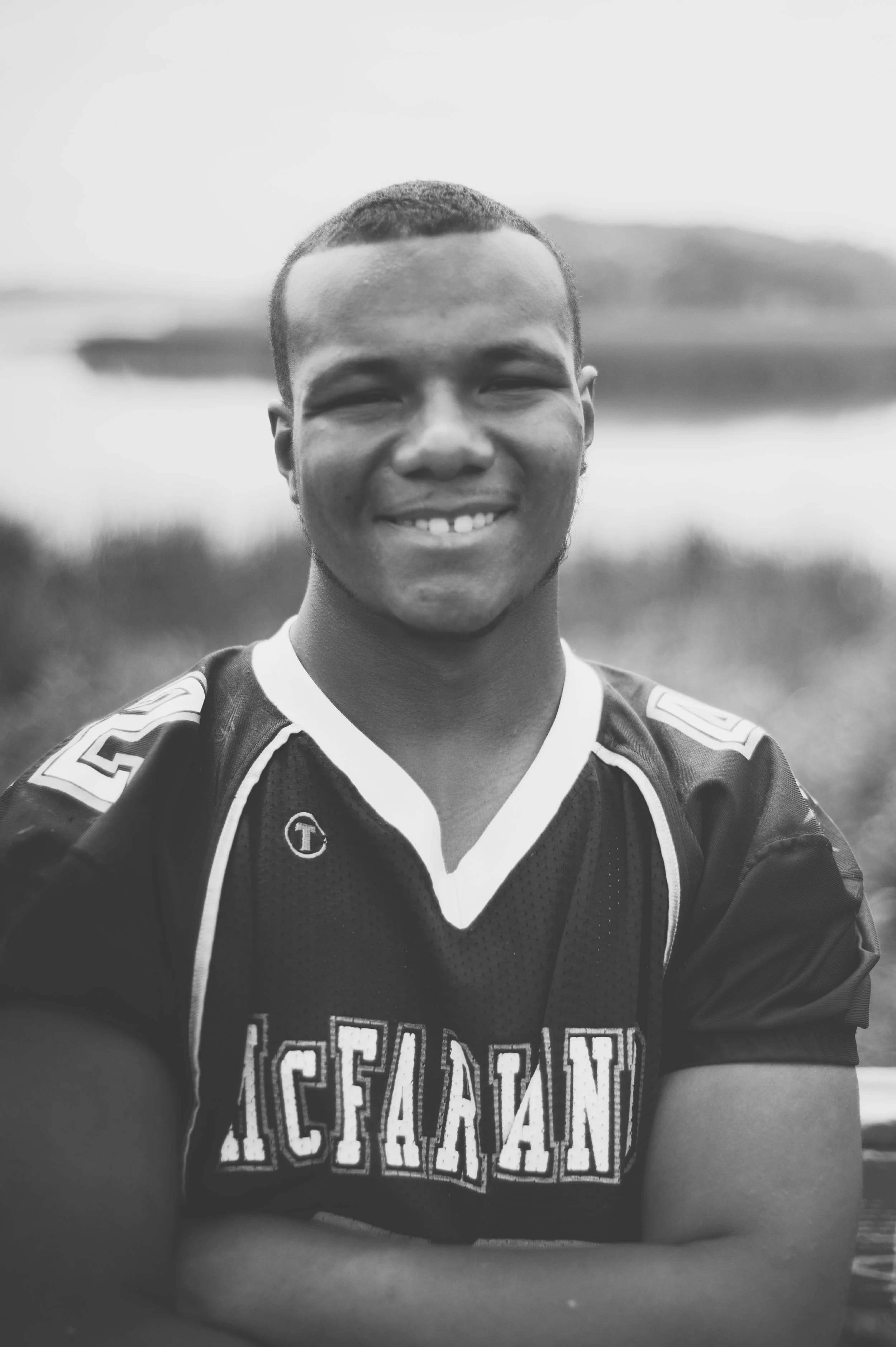 A young man smiling outdoors by a lake, wearing a black sports jersey with the word 'ICEFALAN' on it.
