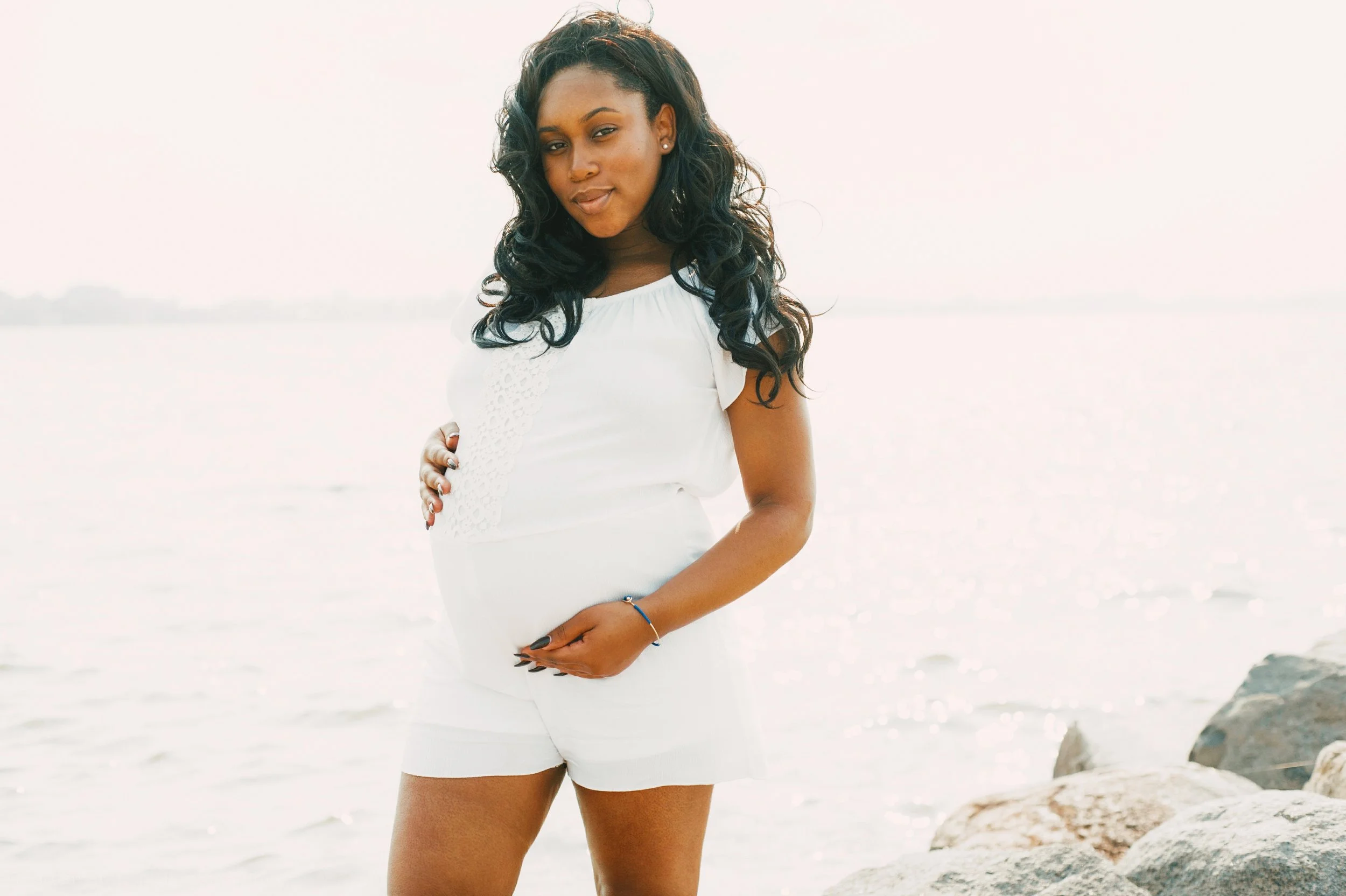 A pregnant woman in a white dress standing on rocks by a body of water, looking at the camera with a soft smile.
