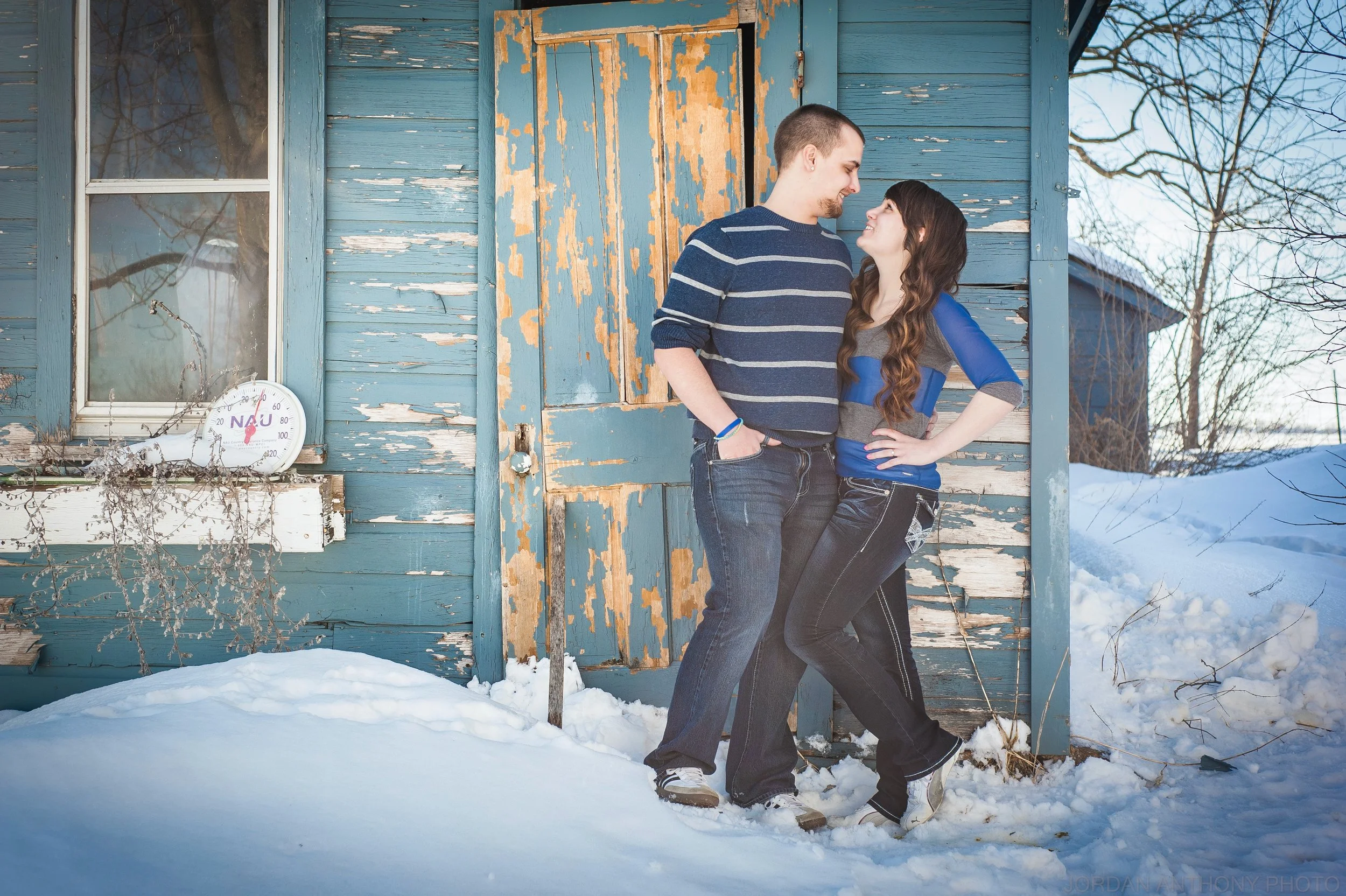 A young couple smiling and standing close together outside a weathered blue wooden house with peeling paint in a snowy landscape.