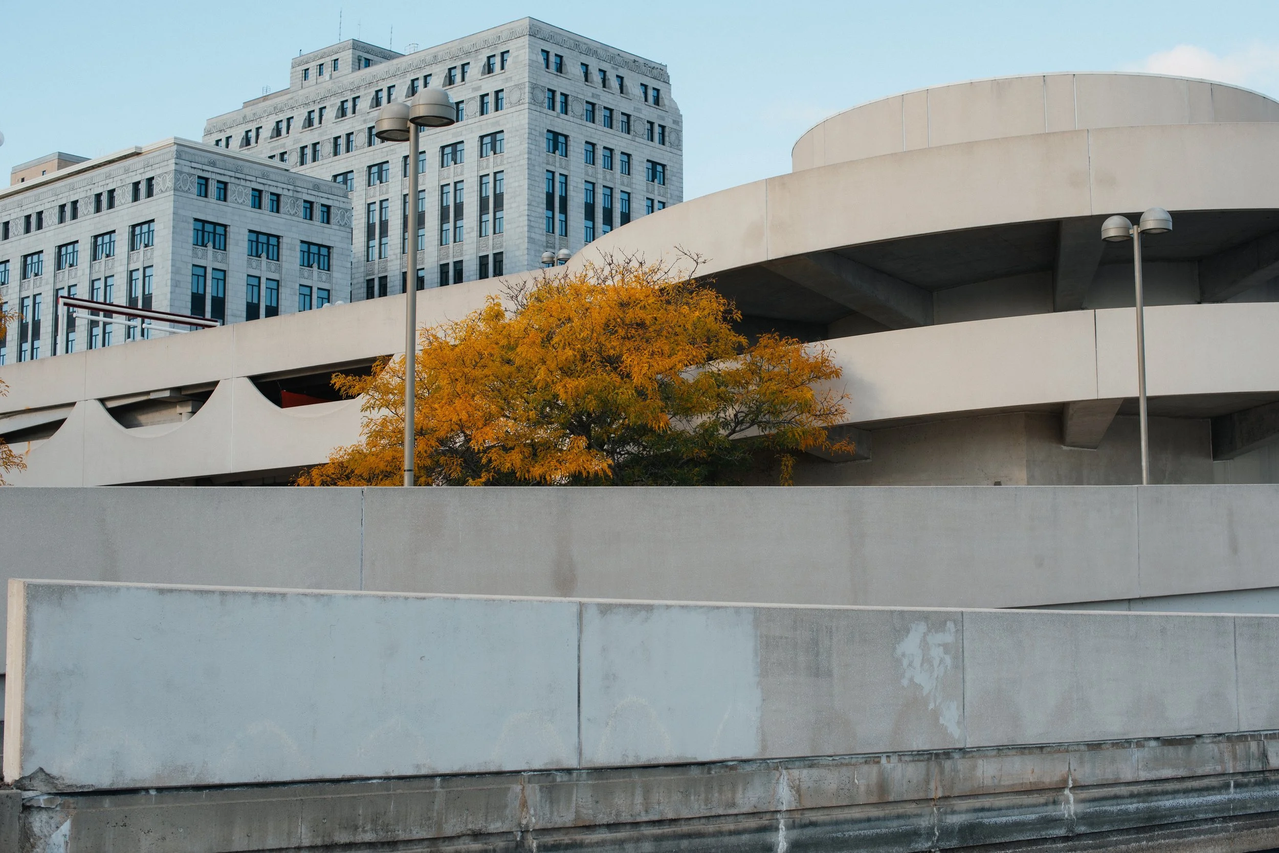 Concrete parking structure with a tree showing orange autumn leaves, and tall office buildings in the background under a partly cloudy sky.