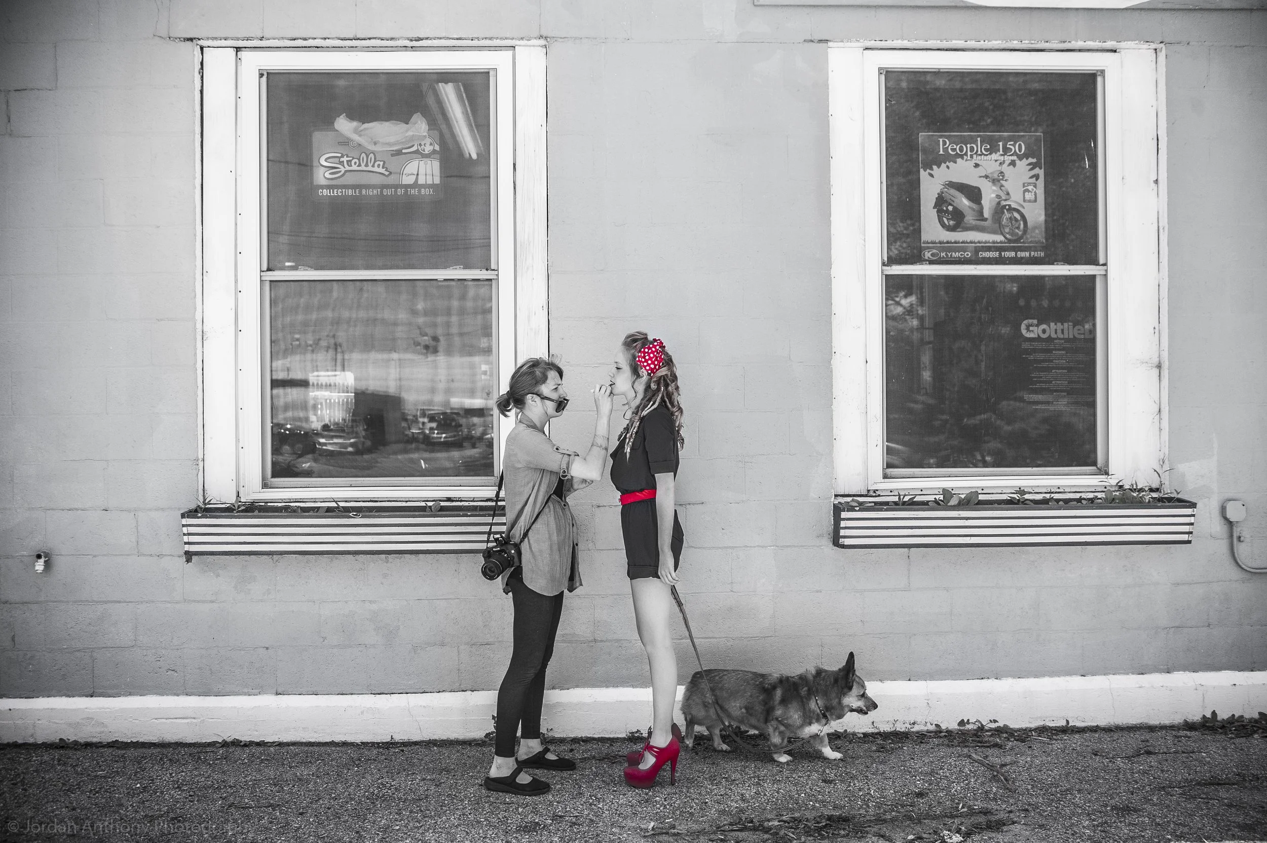 A black-and-white photo of two women and a dog outside a building with two windows. One woman, wearing casual clothes and face mask, is applying makeup to the other woman, who is dressed in a black outfit with red accessories and high heels. The dog 