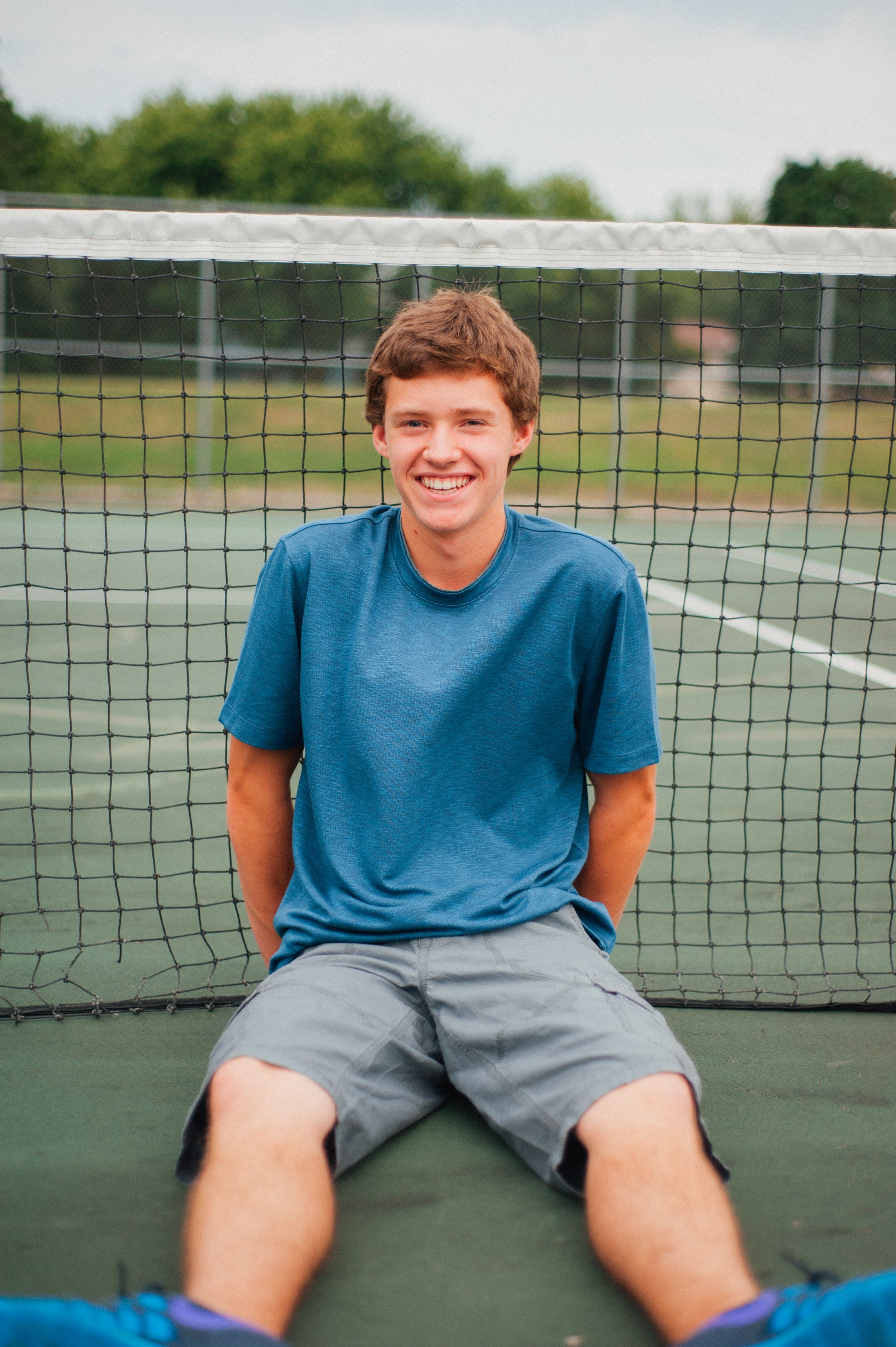 A young man sitting on a tennis court, smiling at the camera, with a tennis net behind him and trees in the background.