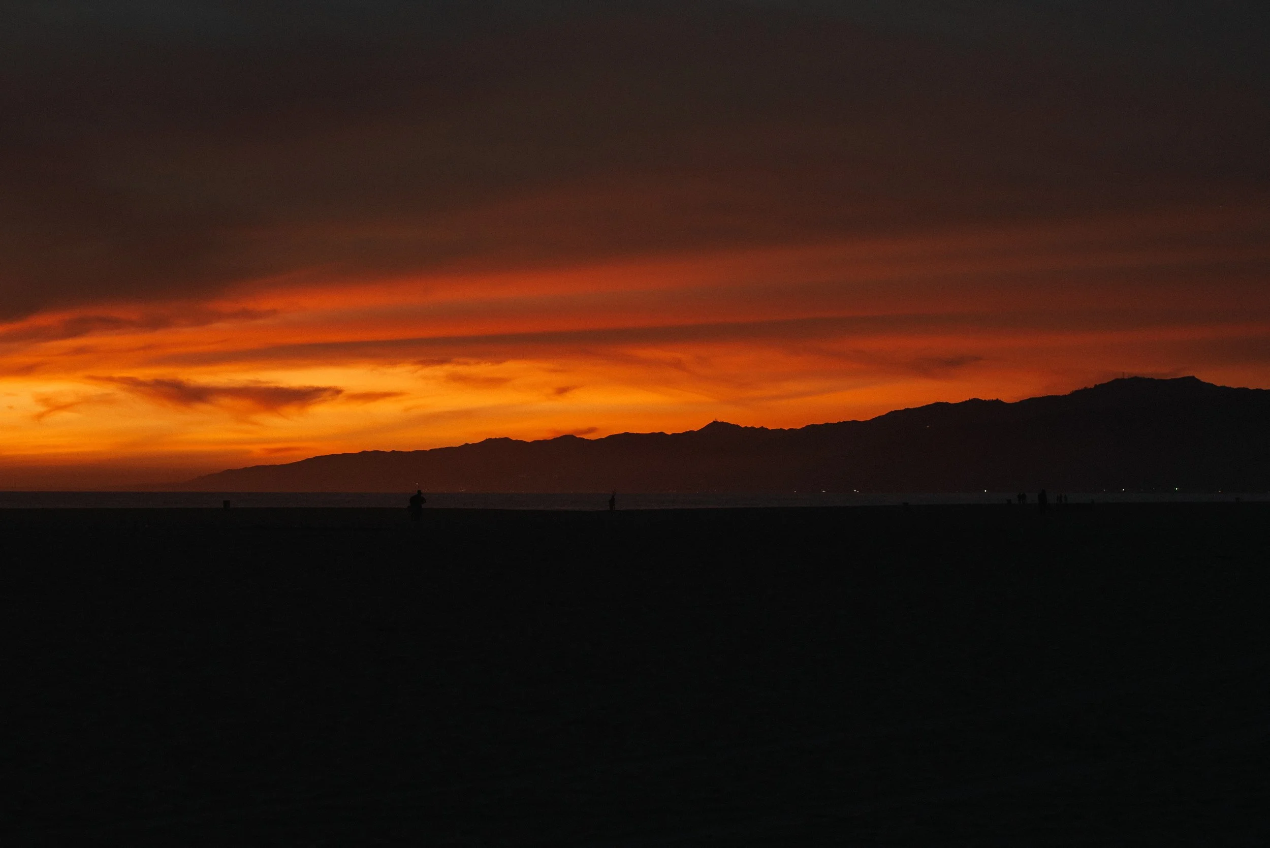 Sunset over the ocean with orange and yellow clouds and silhouettes of people on the beach.