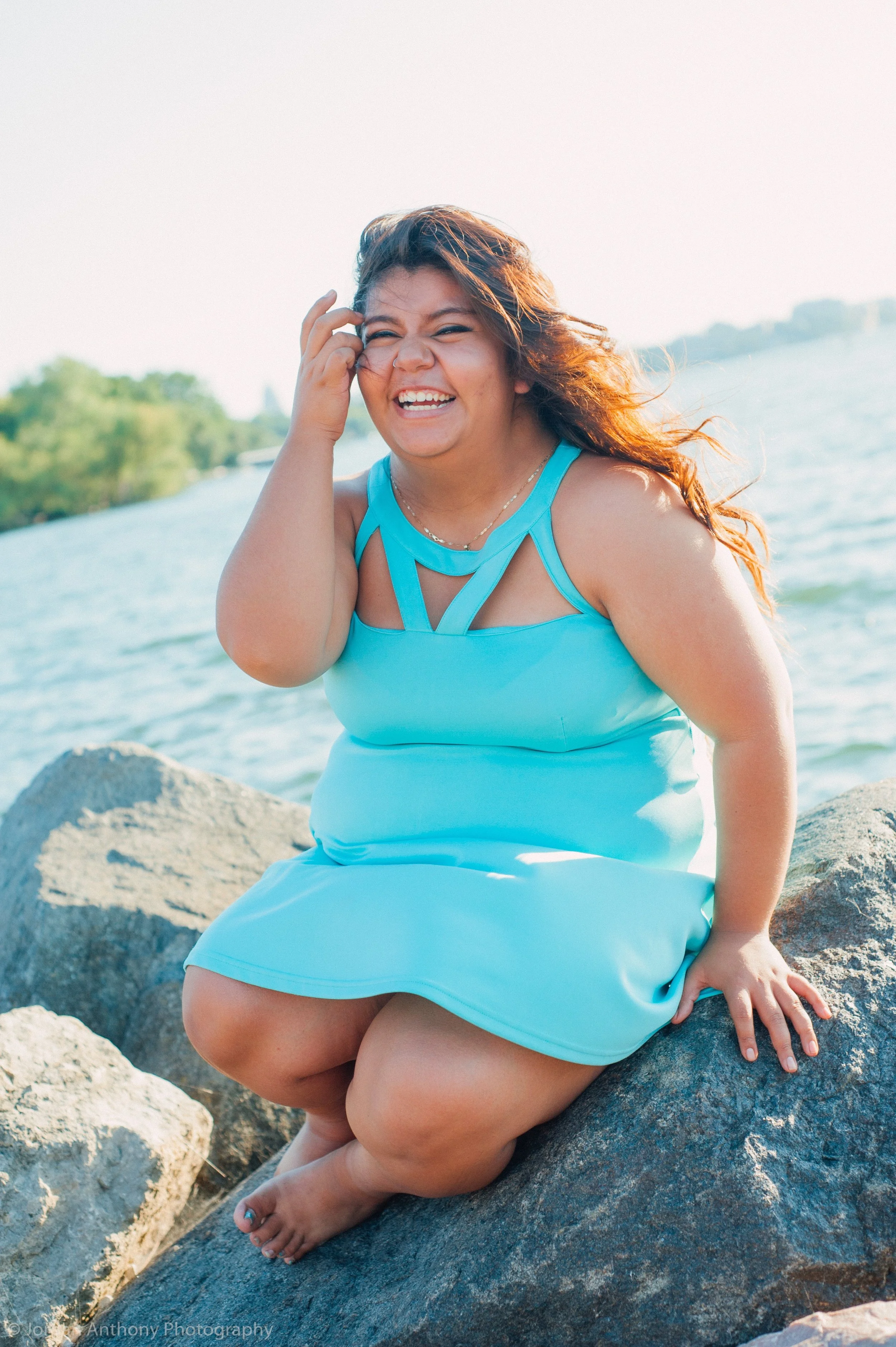 A woman in a light blue dress sitting on large rocks near a body of water, smiling and touching her hair.
