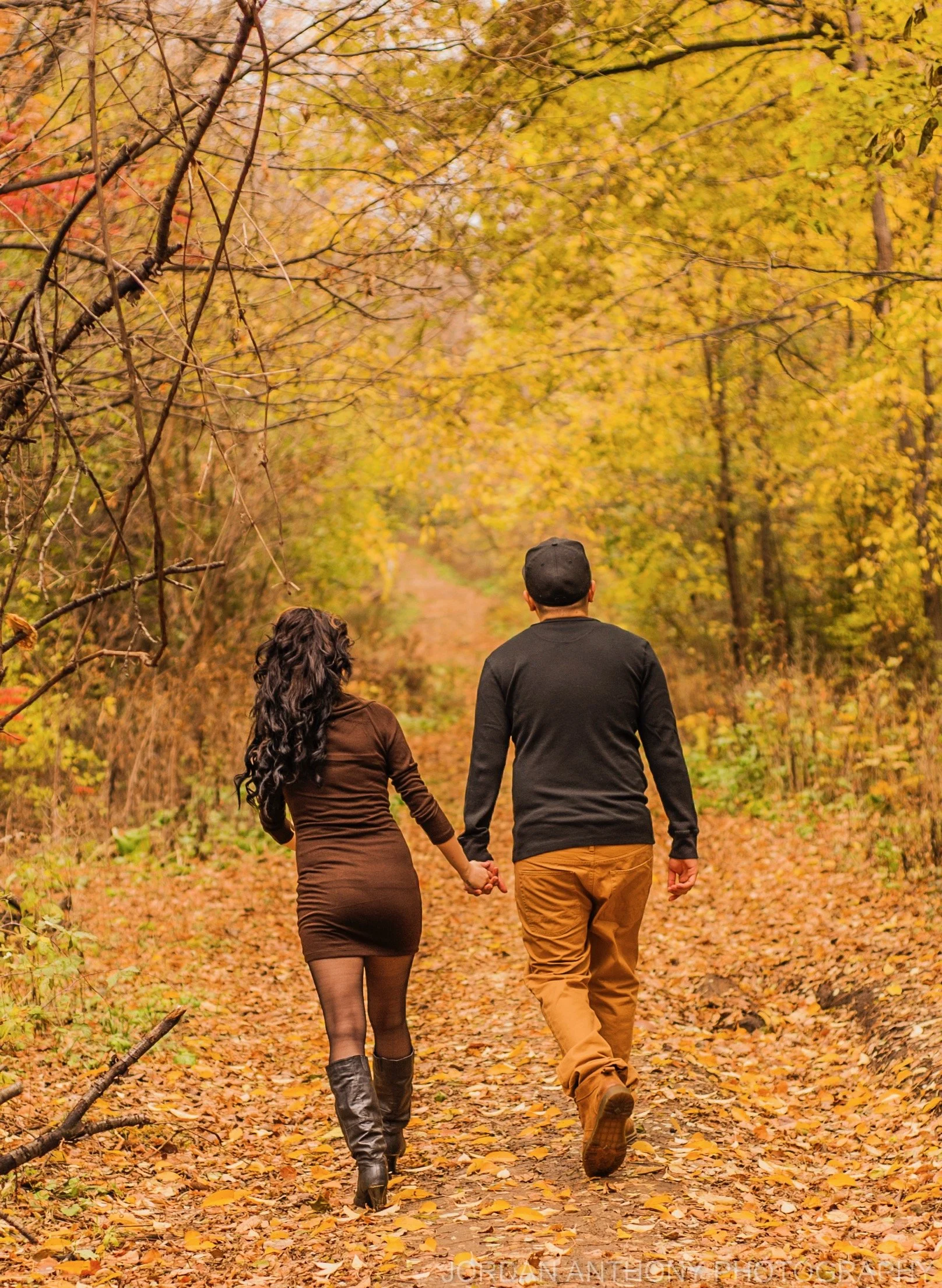 A couple holding hands and walking down an autumn forest trail covered in fallen leaves.