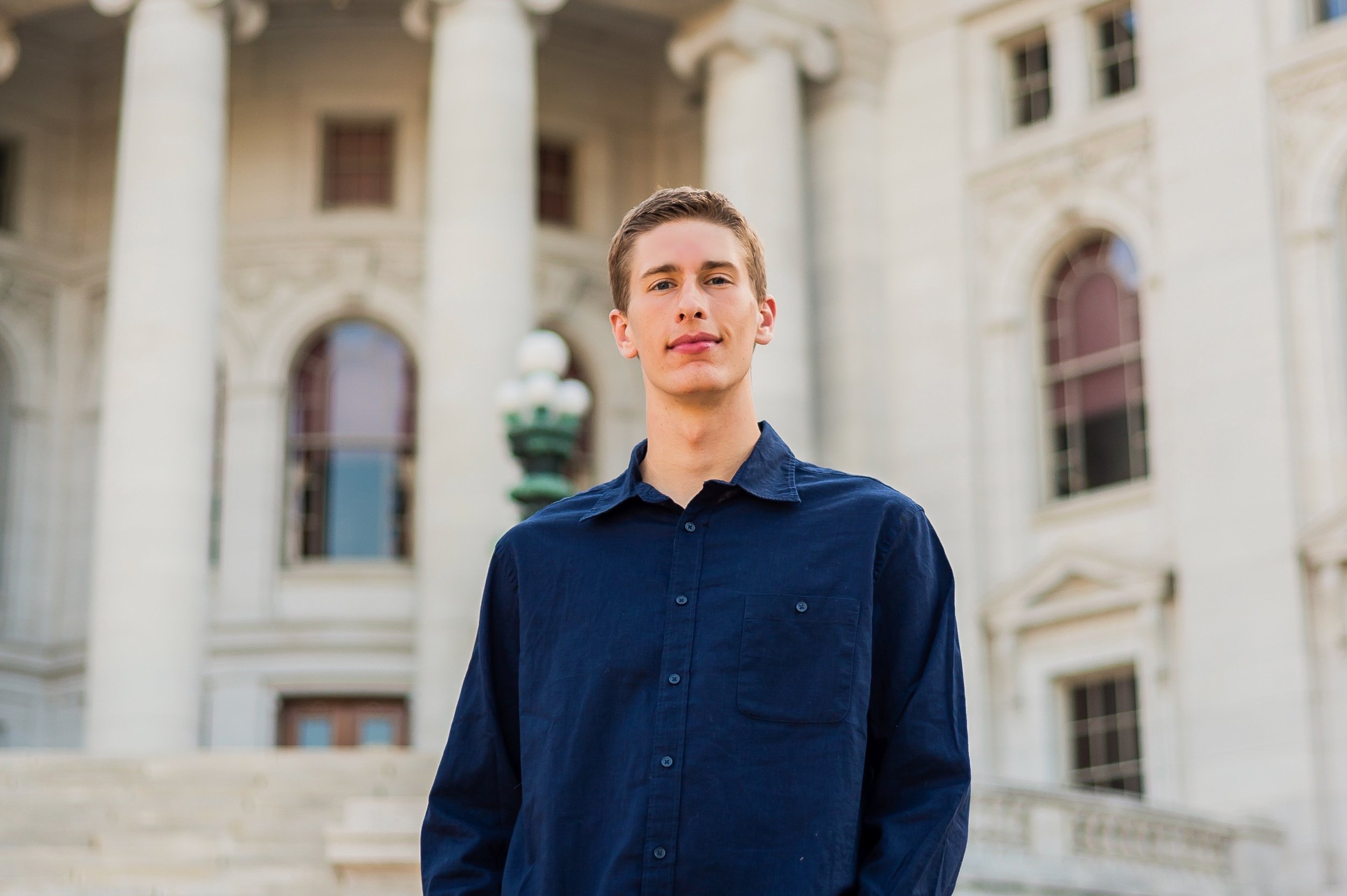 Young man standing in front of a historic government building with large columns and steps, wearing a navy blue shirt.