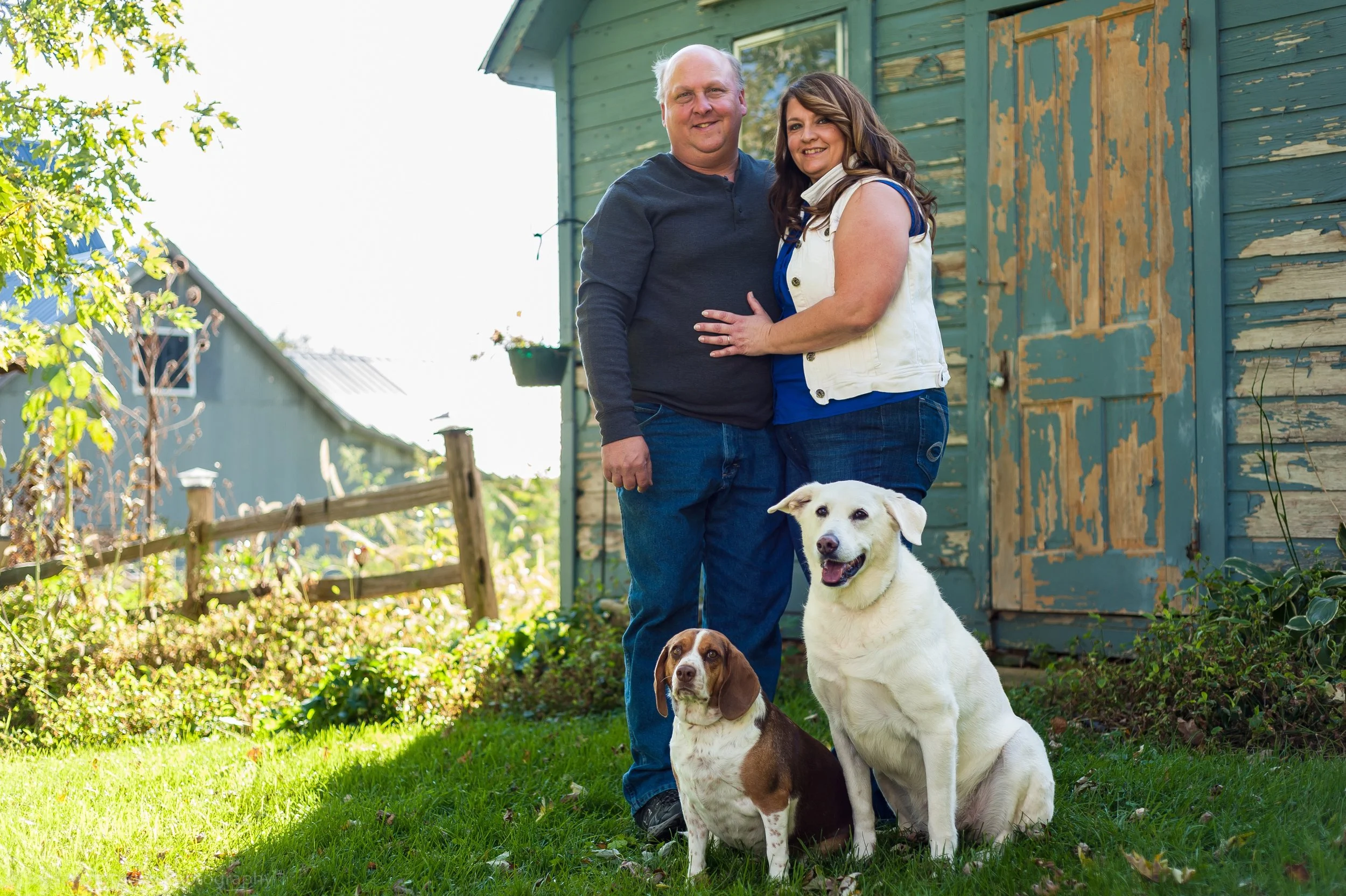 A couple standing outside in front of a wooden shed with two dogs, one sitting and one standing, in the yard on a sunny day.