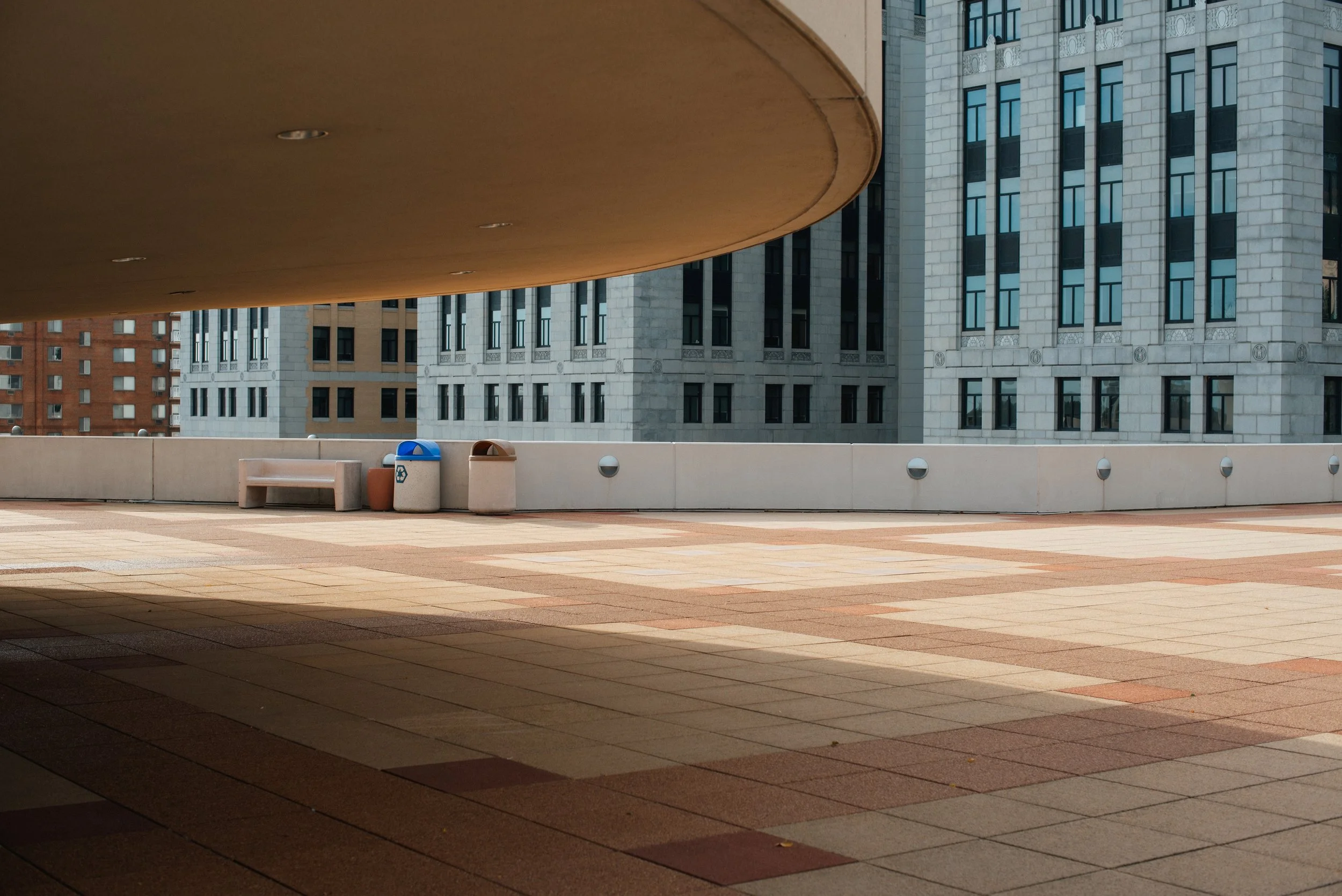 Empty outdoor terrace with a concrete bench and two recycling bins, overlooking tall office buildings.