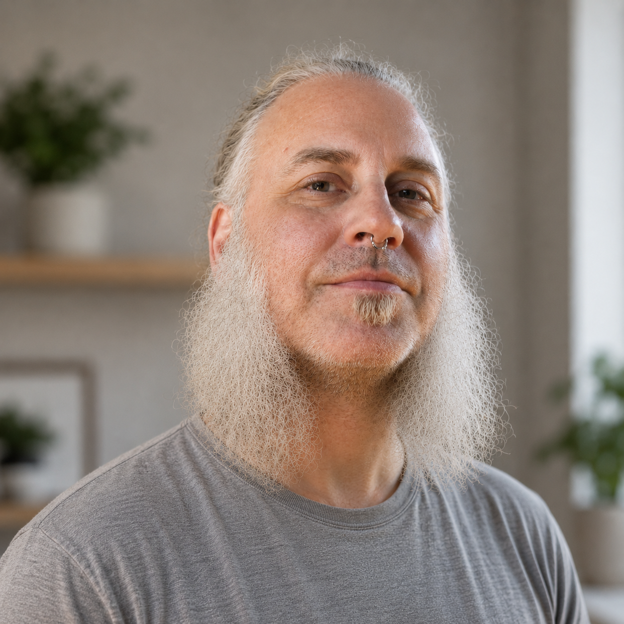 Close-up of a smiling man with a long, gray beard and a septum piercing, with a dark background.