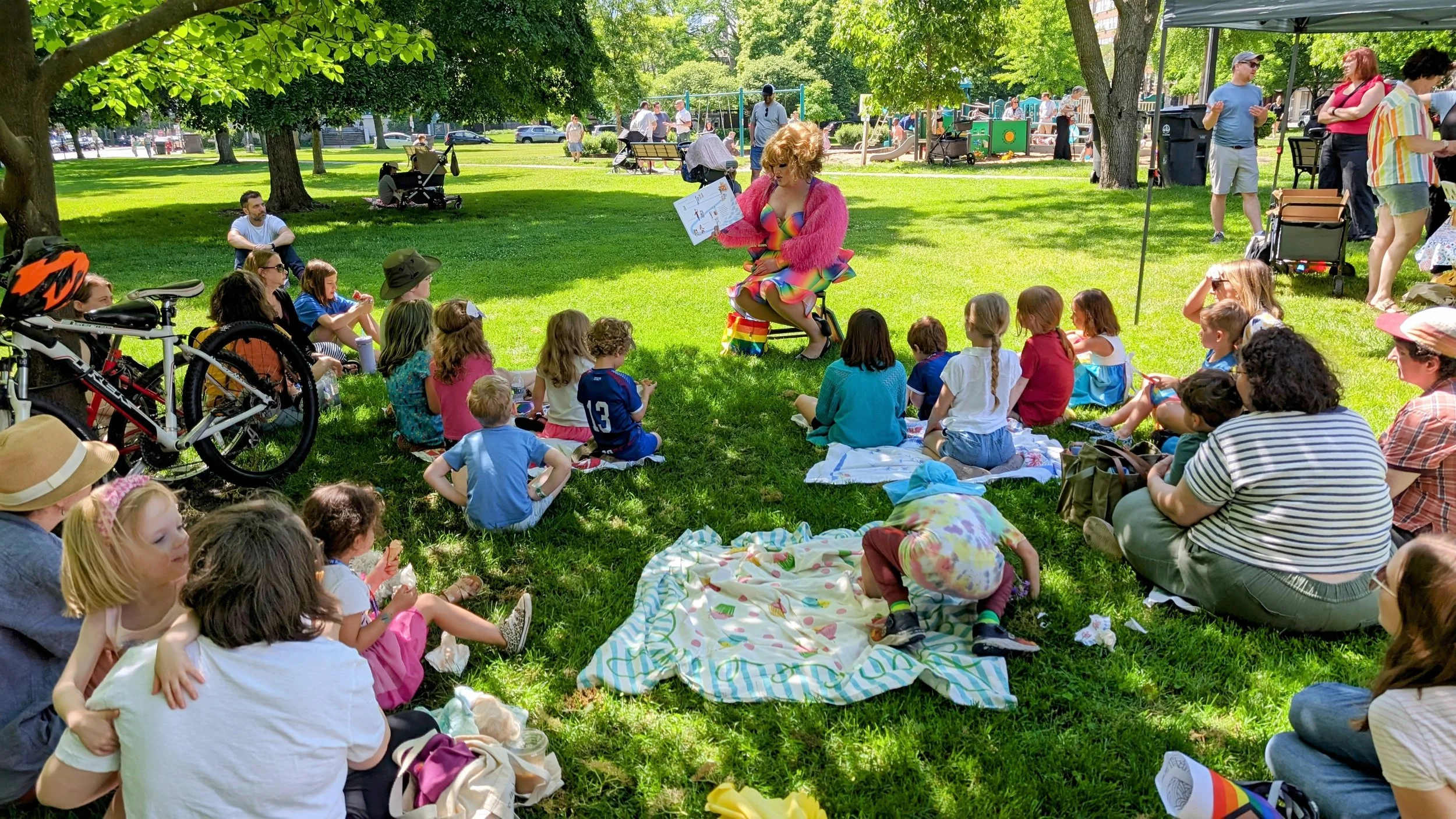 A group of children and adults gathered in a park, watching a performer dressed in colorful rainbow attire, sitting on a stool and reading a book. The audience is seated on the grass under trees, with some on blankets, and includes families with stro