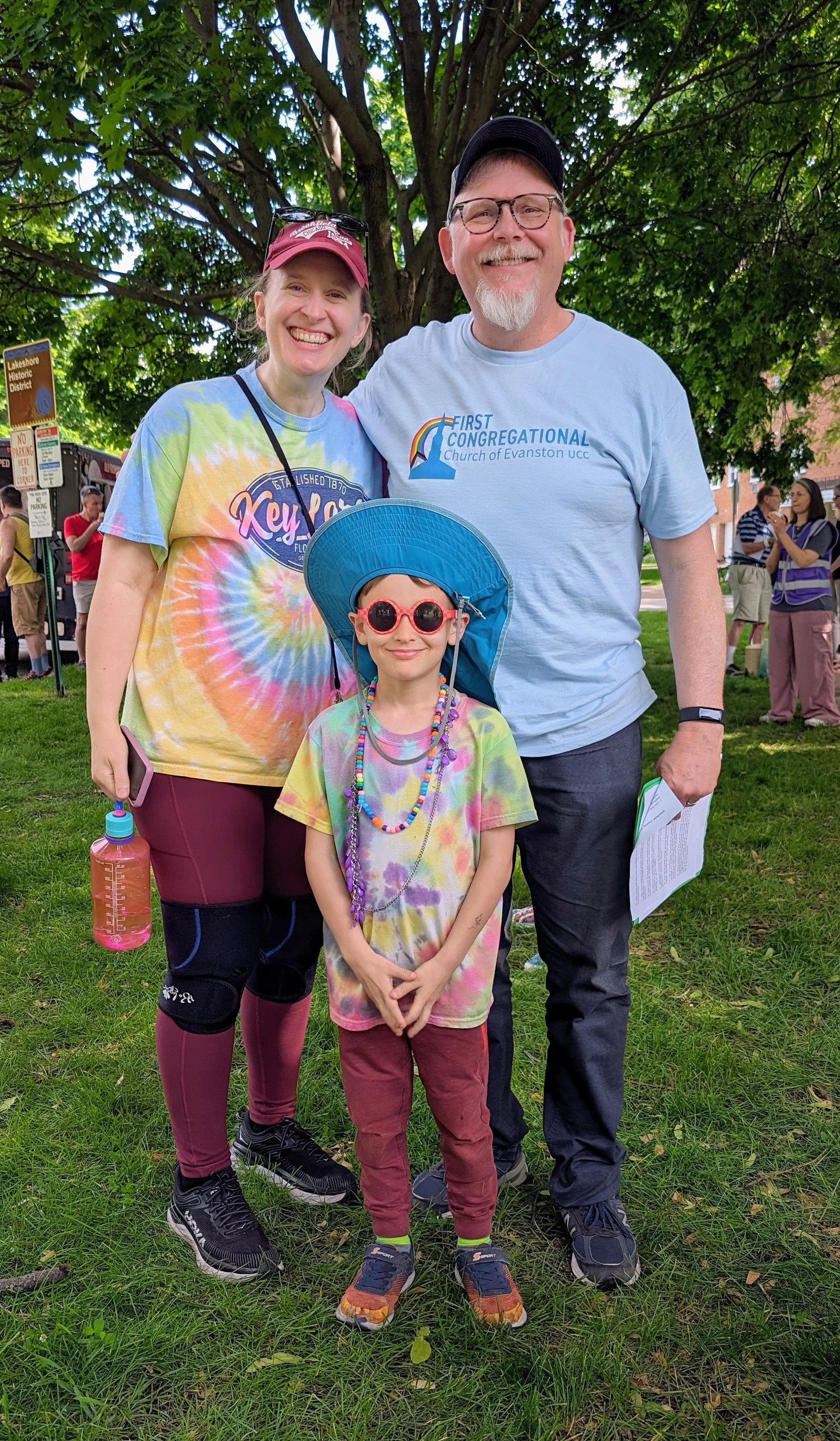 A family of three smiling at an outdoor event with a large tree and other people in the background. The mother wears a colorful tie-dye shirt, the father wears a light blue T-shirt and glasses, and the child wears a tie-dye shirt, a blue sun hat, and