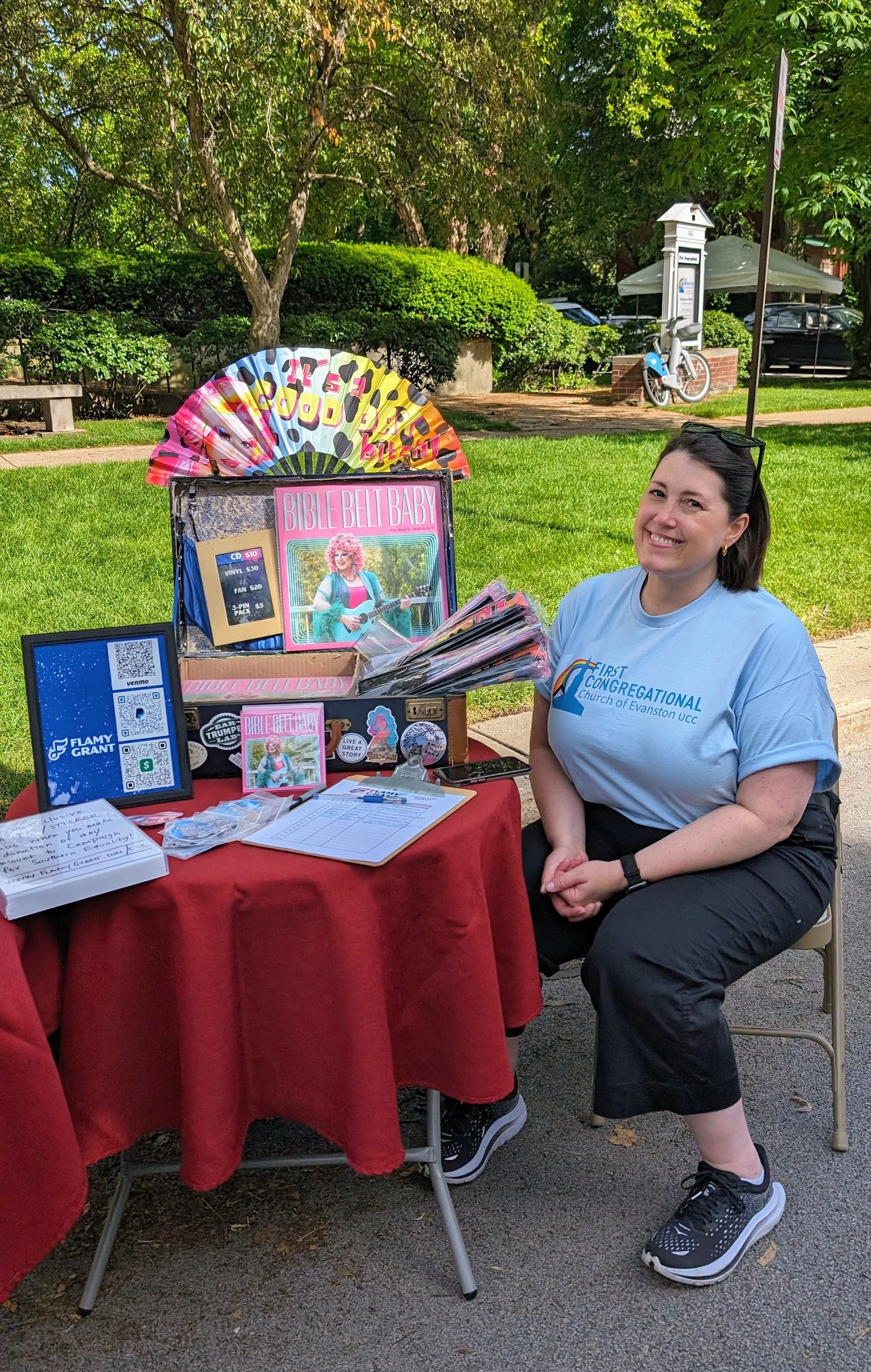 A woman sitting at a table outdoors at a church event, with various items on display including CDs, books, and a sign, with trees and a bike in the background.