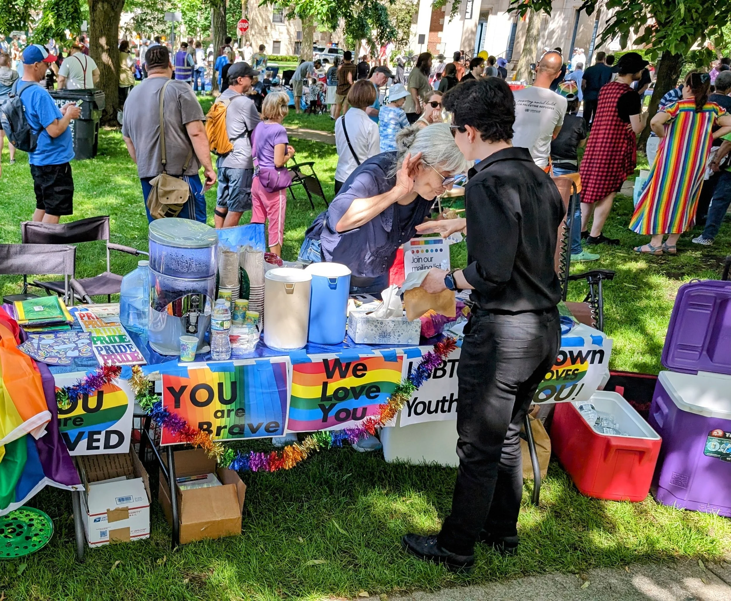 A crowd gathered at an outdoor event in a park; a table decorated with pride banners and rainbow decorations serves attendees, with two women engaging in conversation at the table.
