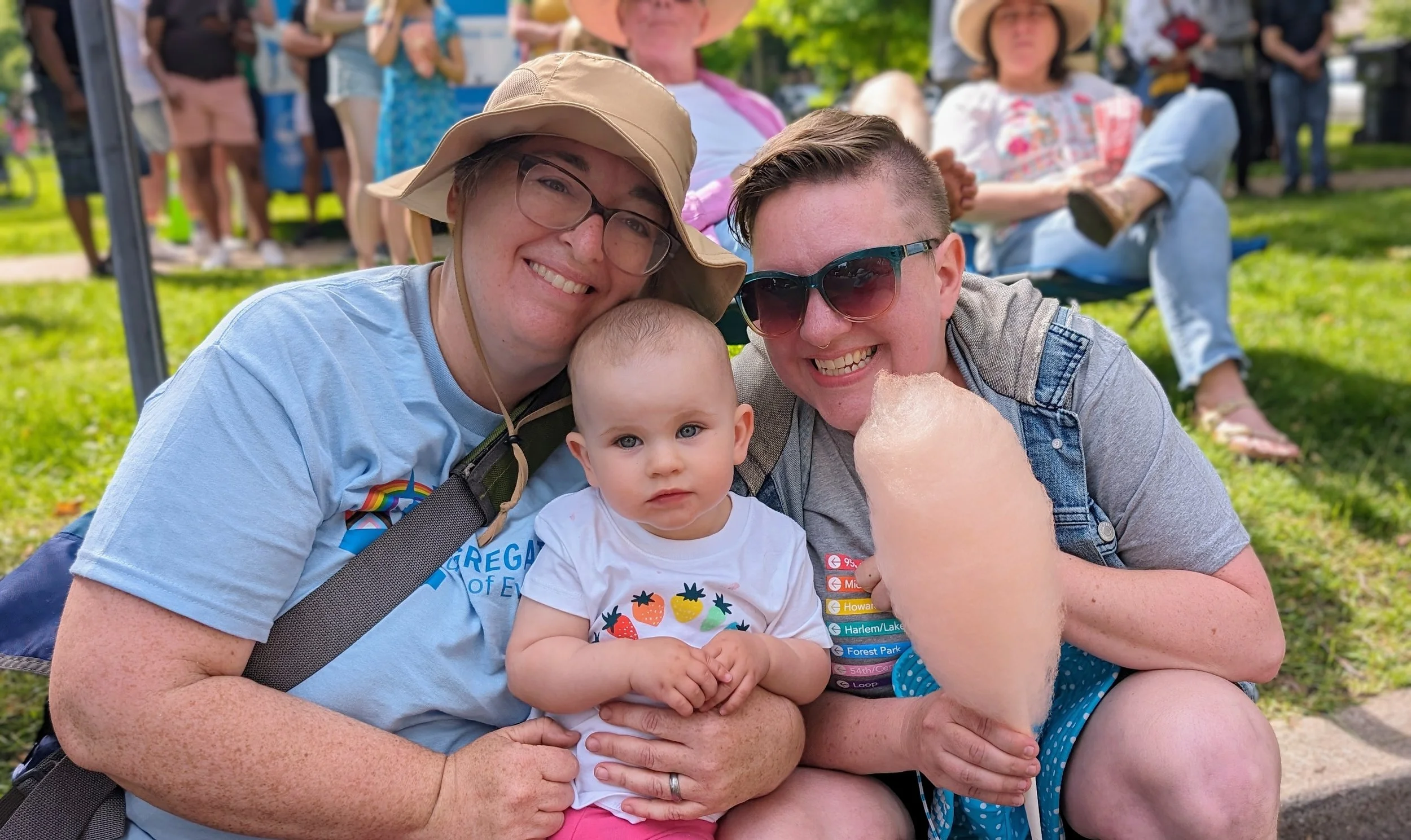 Three people, including a baby girl, a woman with glasses and a man with sunglasses, smiling and posing for a photo outdoors at a festival or fair. The woman is wearing a beige hat and the man is holding cotton candy. In the background, there are oth