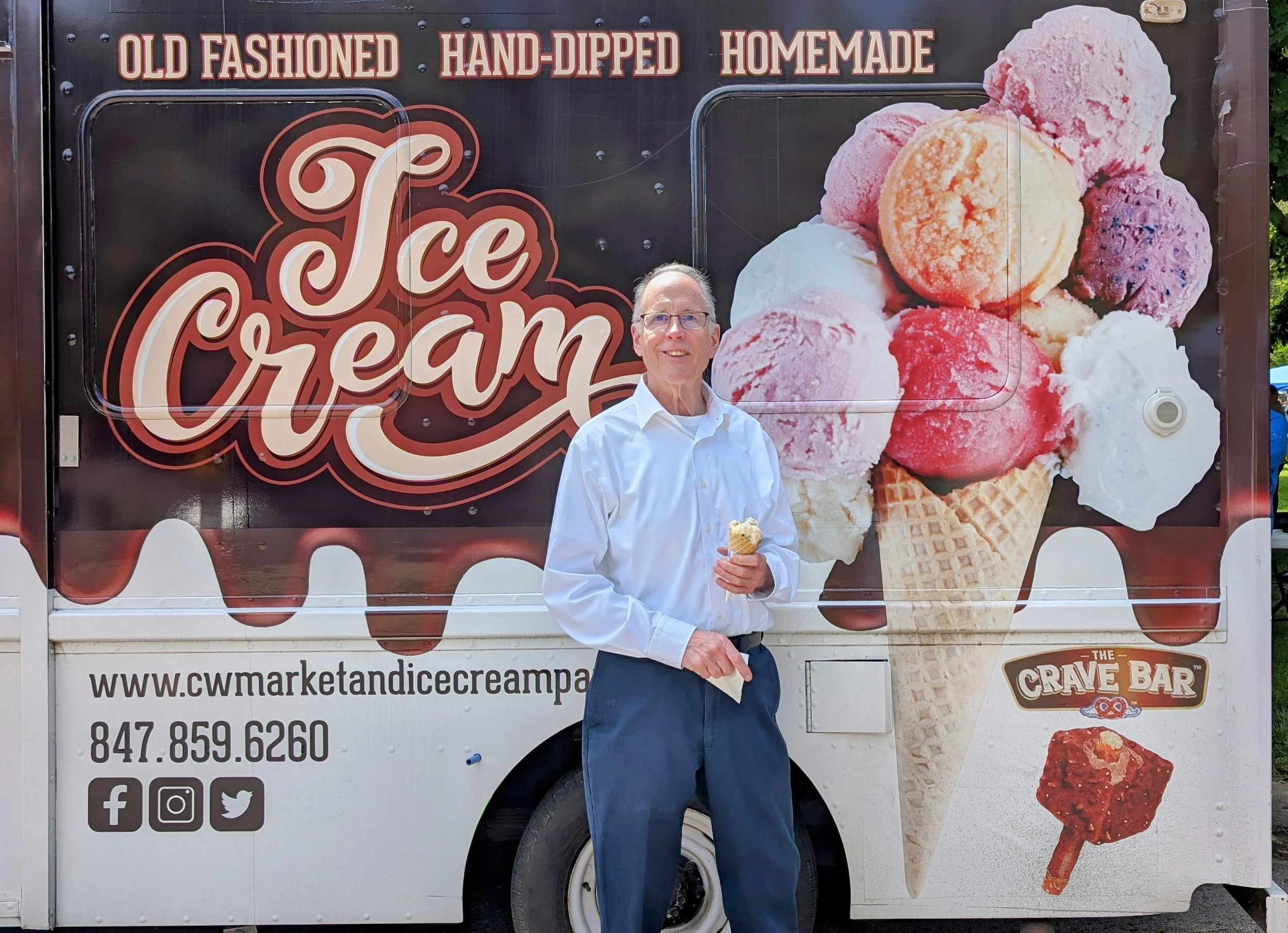 A man in a white shirt and dark pants holding an ice cream cone standing in front of an ice cream truck with colorful ice cream images and the words "Old Fashioned Hand-Dipped Homemade Ice Cream."