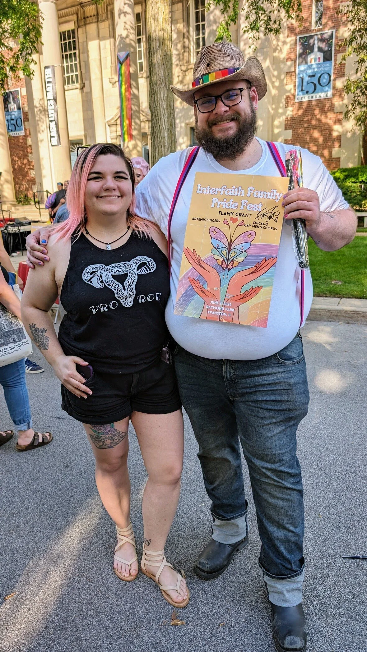 A smiling woman with pink hair and tattoos standing next to a bearded man wearing glasses and a cowboy hat at Pride fest. The man is holding a colorful flyer for Interfaith Family Pride Fest, June 2024, Raymon Park, Evanston, Illinois.
