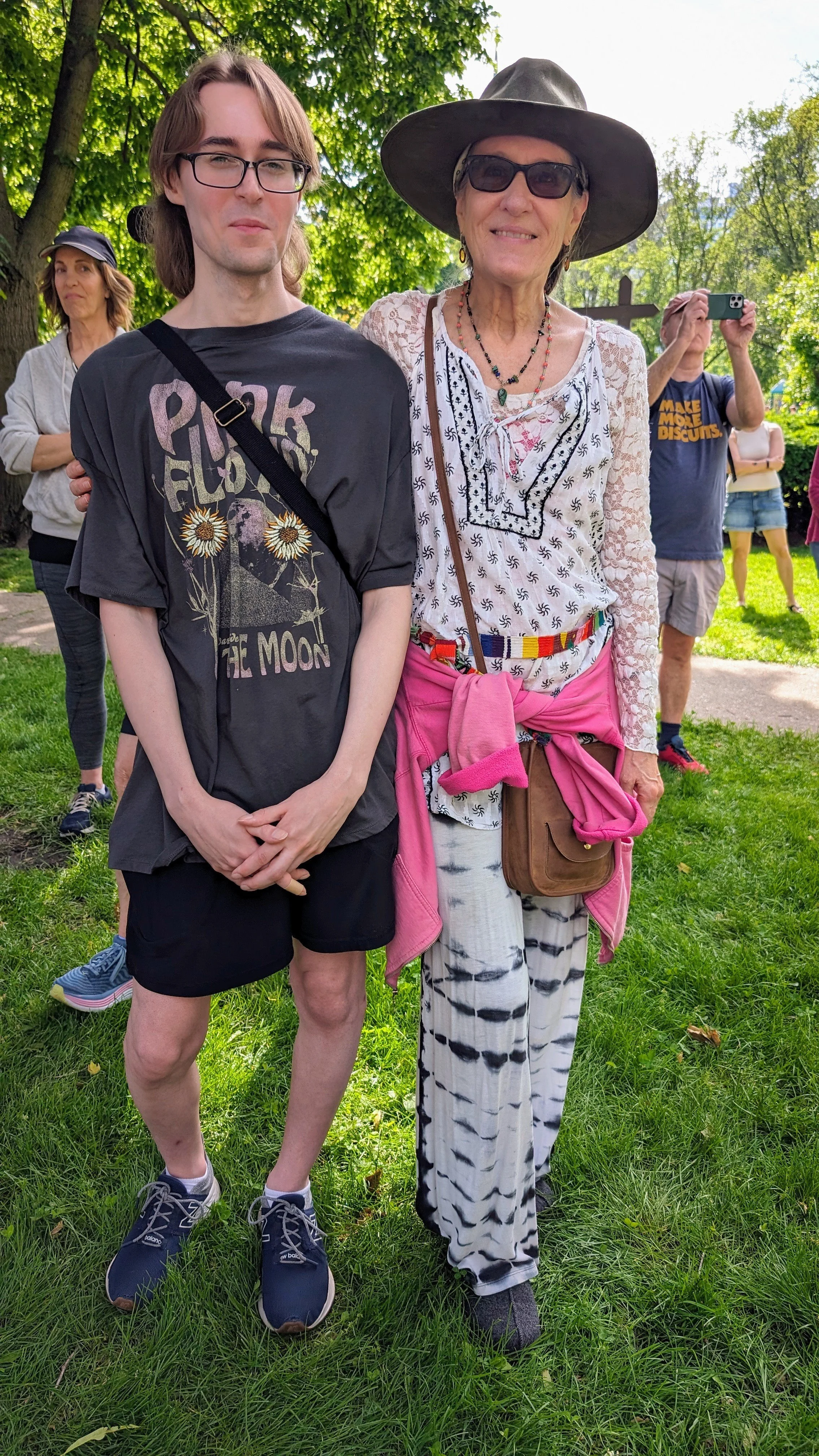 A young man and an older woman standing together outdoors on a sunny day, with other people in the background. The young man is wearing a graphic T-shirt, black shorts, and sneakers, with glasses. The older woman is wearing a large sunhat, sunglasses