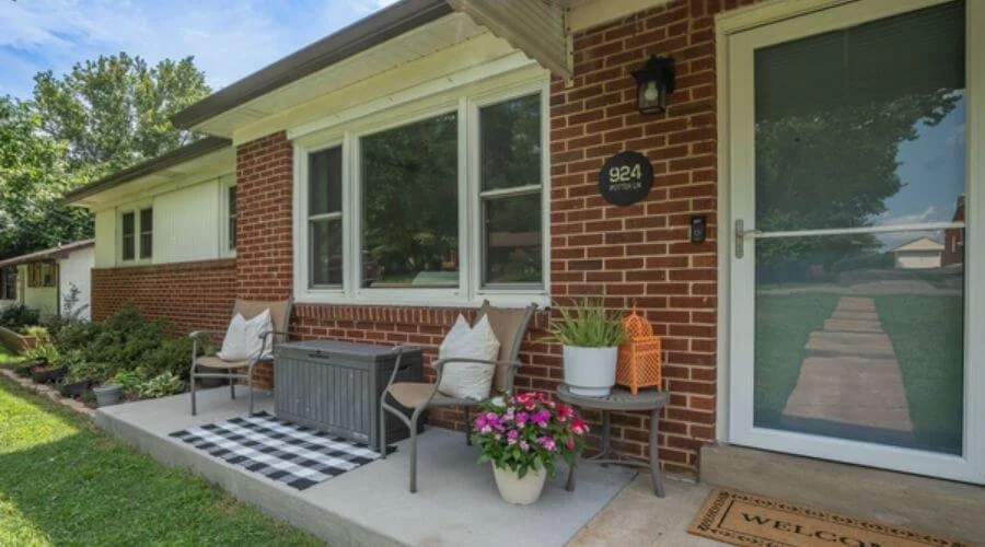 Front porch view of a single-level, red-bricked home with minimalist outdoor furniture and large front window.