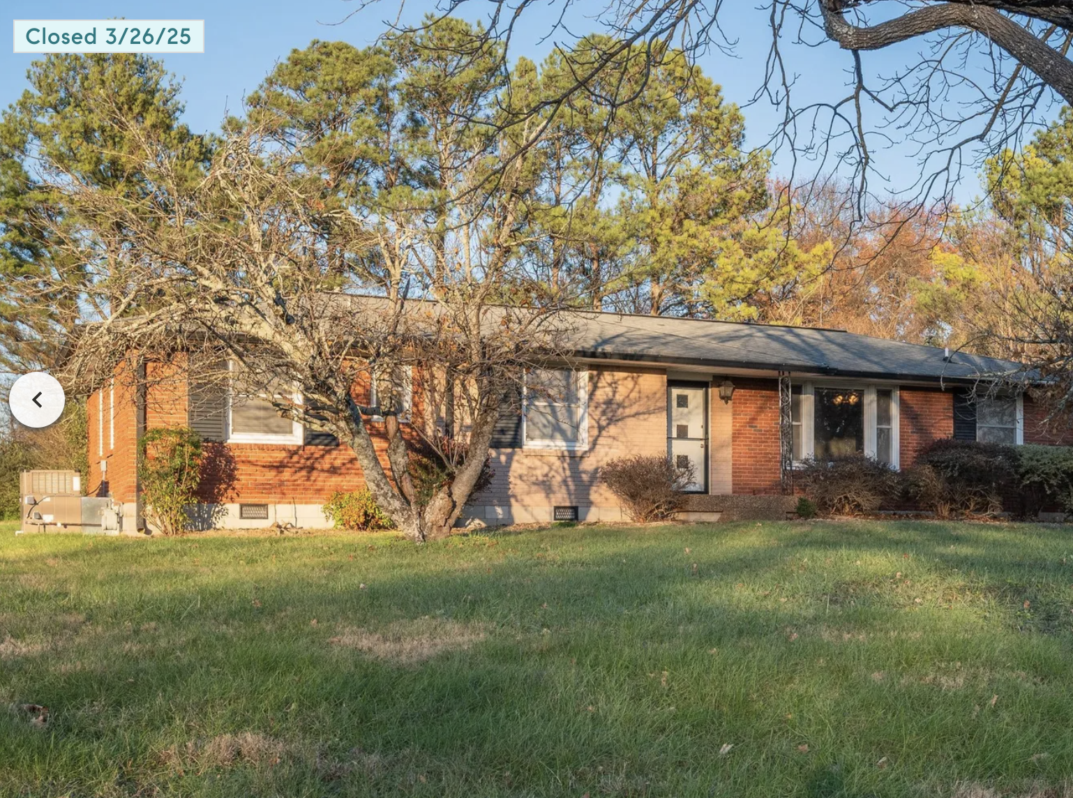 A single-story brick house with a front lawn, leafless trees, and bushes, with the house doors and windows visible, during late afternoon sunlight.