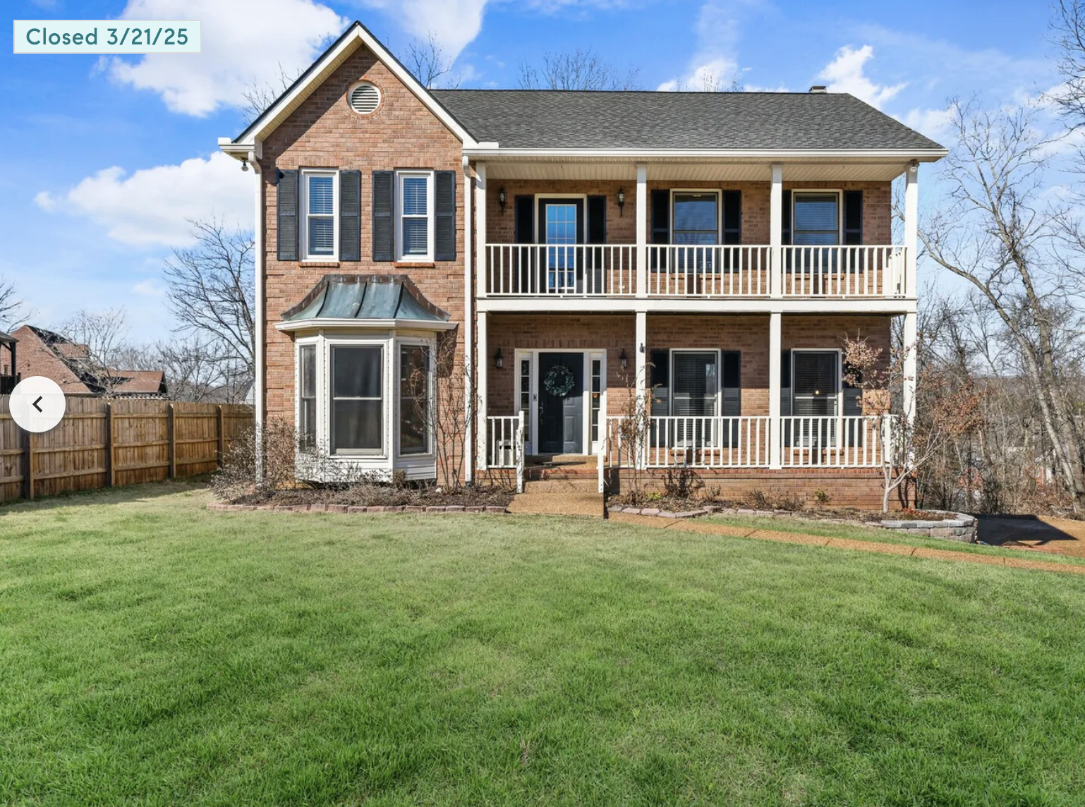 A two-story brick house with white railings on balconies, black shutters on windows, a small front garden with bushes, and a green lawn, under a partly cloudy sky.