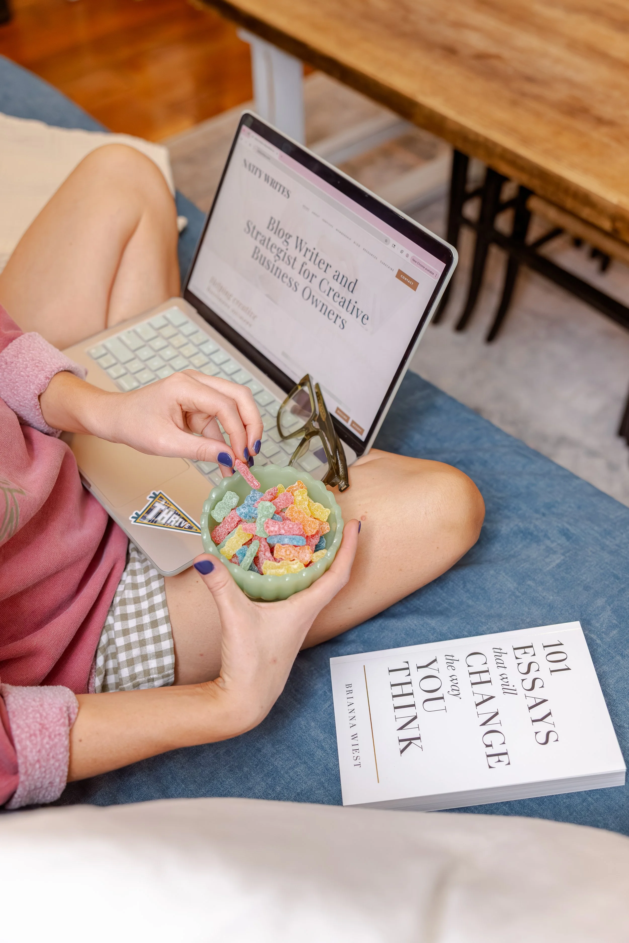 Natty, blogging and content strategist, sitting on a blue couch holding a bowl of colorful gummy bears, with a laptop on their lap open to a website about blogging strategies for business owners, and a book on table.