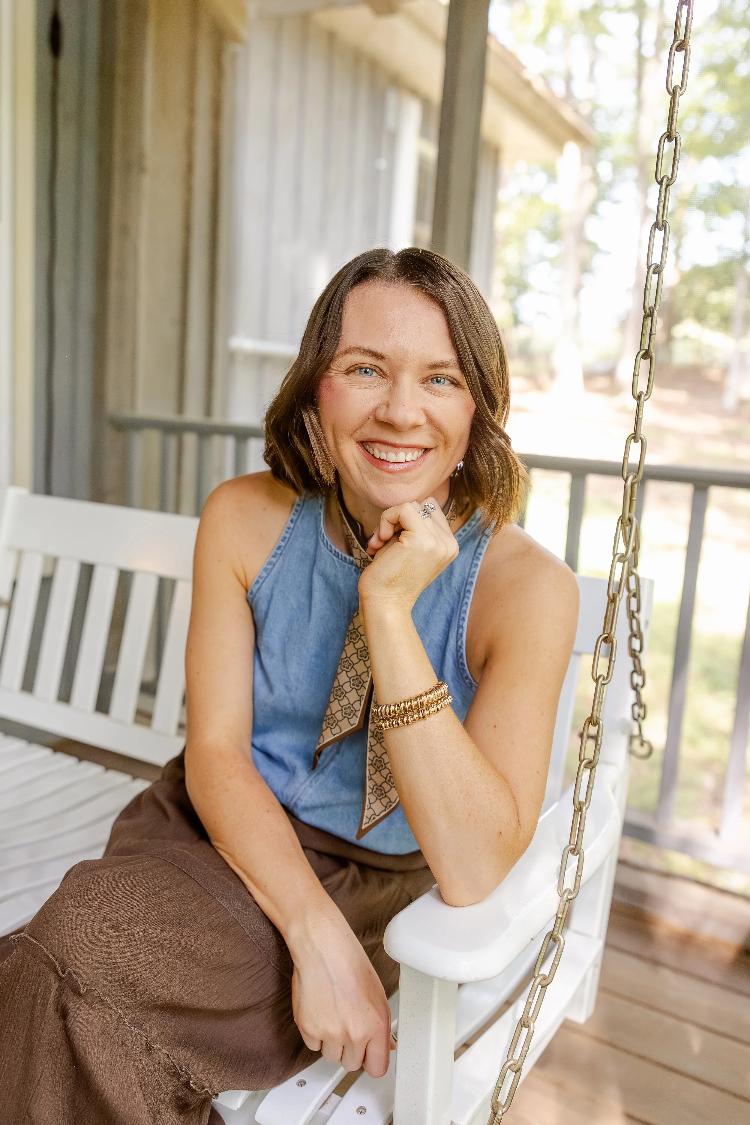Natty Writes wearing a sleeveless denim top, sitting on a white porch swing with chains, on a wooden porch with a background of trees and sunlight.